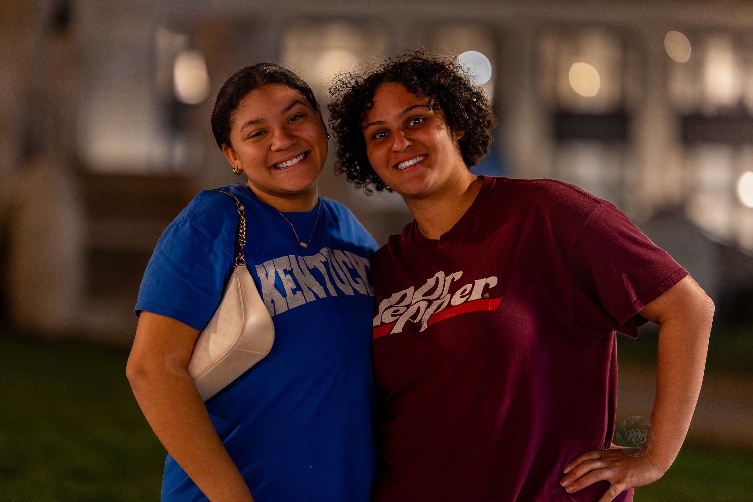 Two women smiling and posing together outdoors at night, with blurred lights in the background.