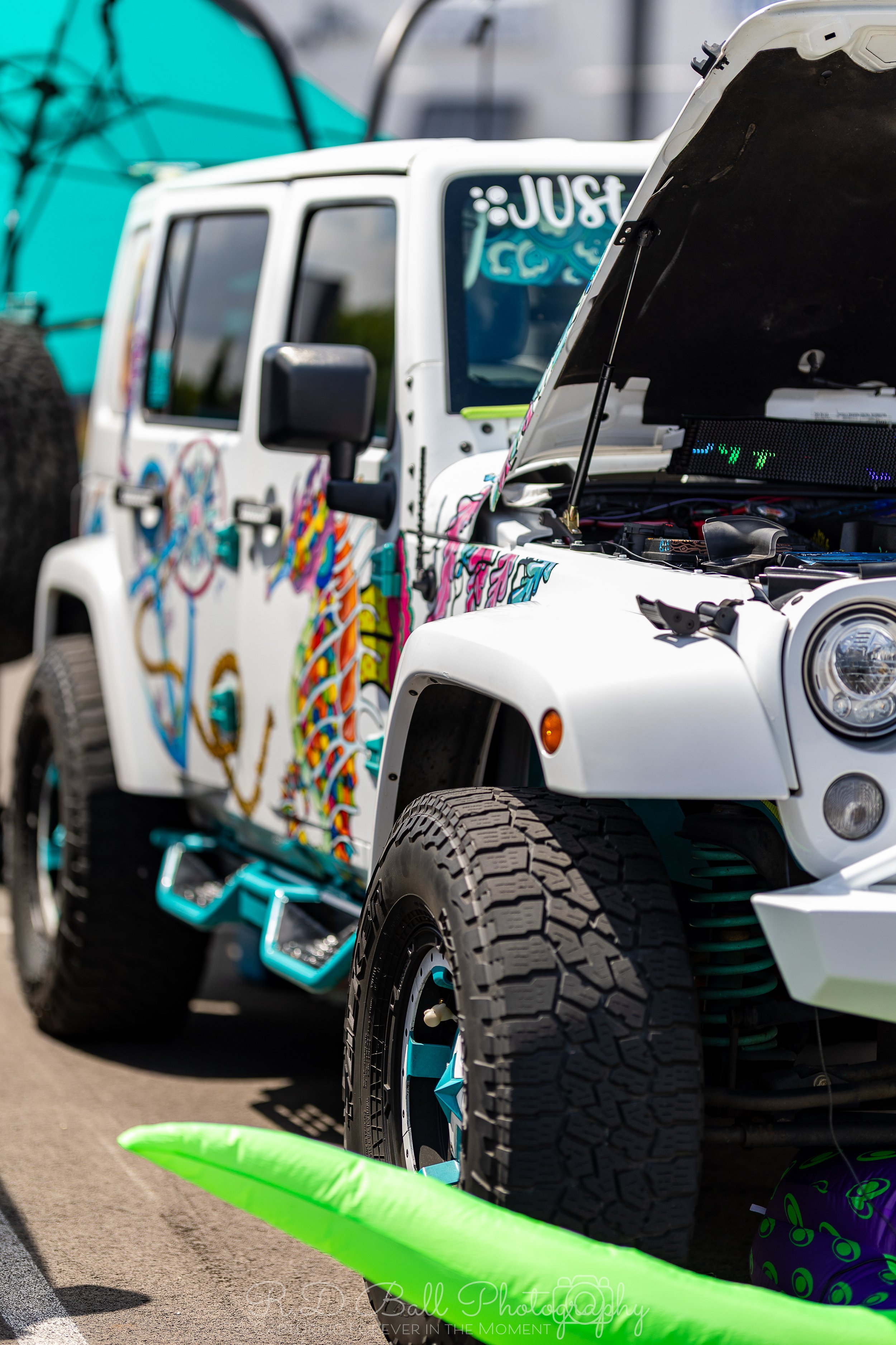 A white Jeep with colorful graffiti-style decorations on the side and hood, with its hood open, parked outdoors during daytime.