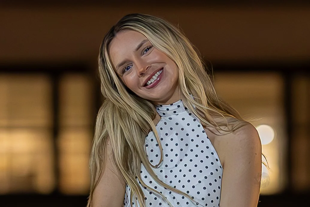 A smiling blonde woman with long hair wearing a sleeveless polka dot top.