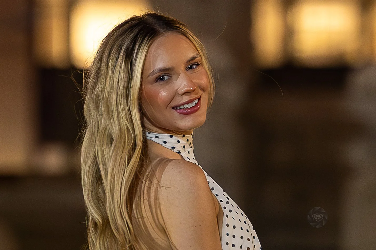 A woman with long blonde hair, smiling and looking over her shoulder. She is wearing a sleeveless white dress with black polka dots. The background is blurred with warm lighting.
