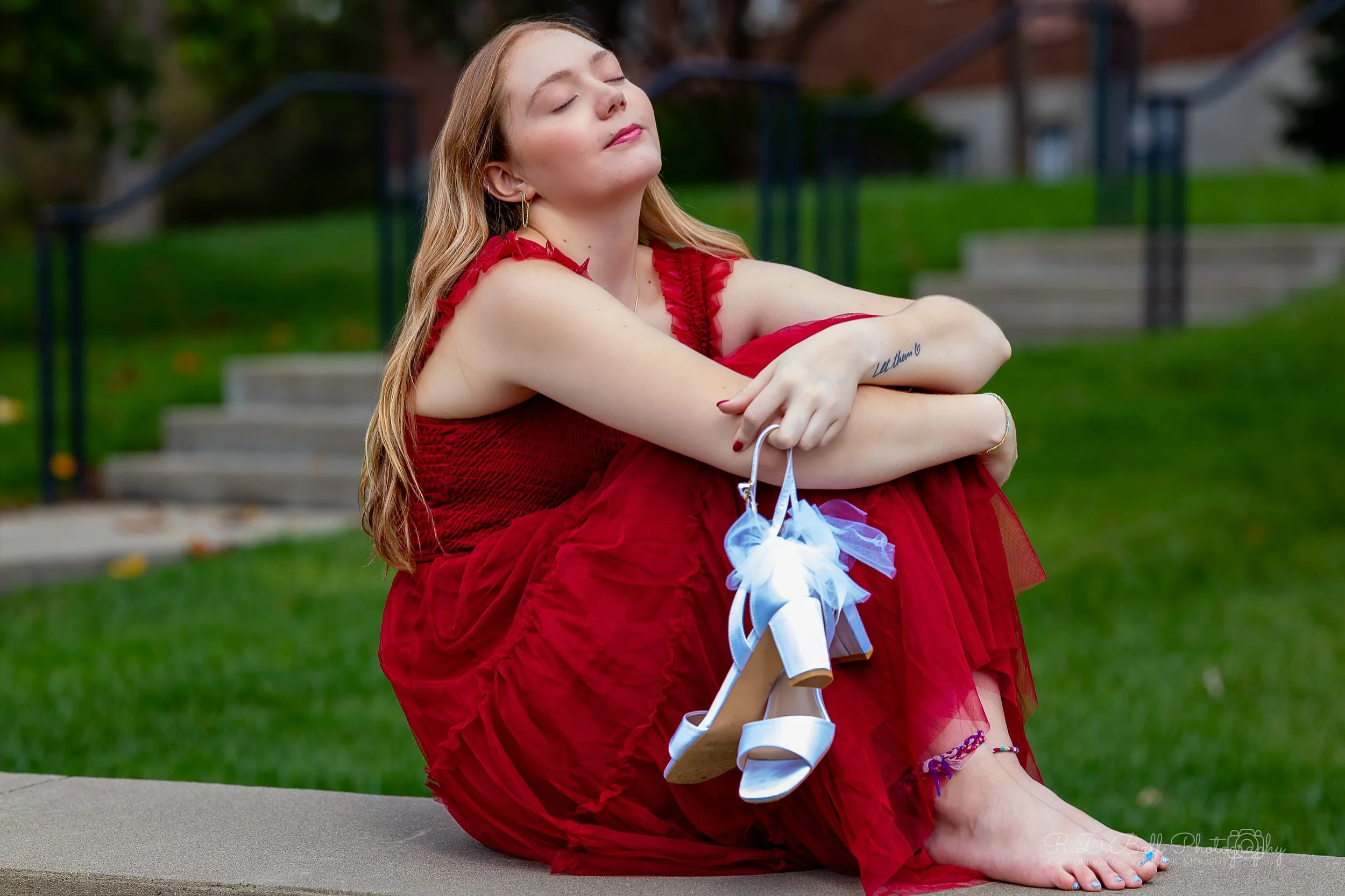 A young woman with long blonde hair sitting on a concrete step outdoors, wearing a red dress and holding white high-heeled shoes, with her eyes closed and a relaxed expression.