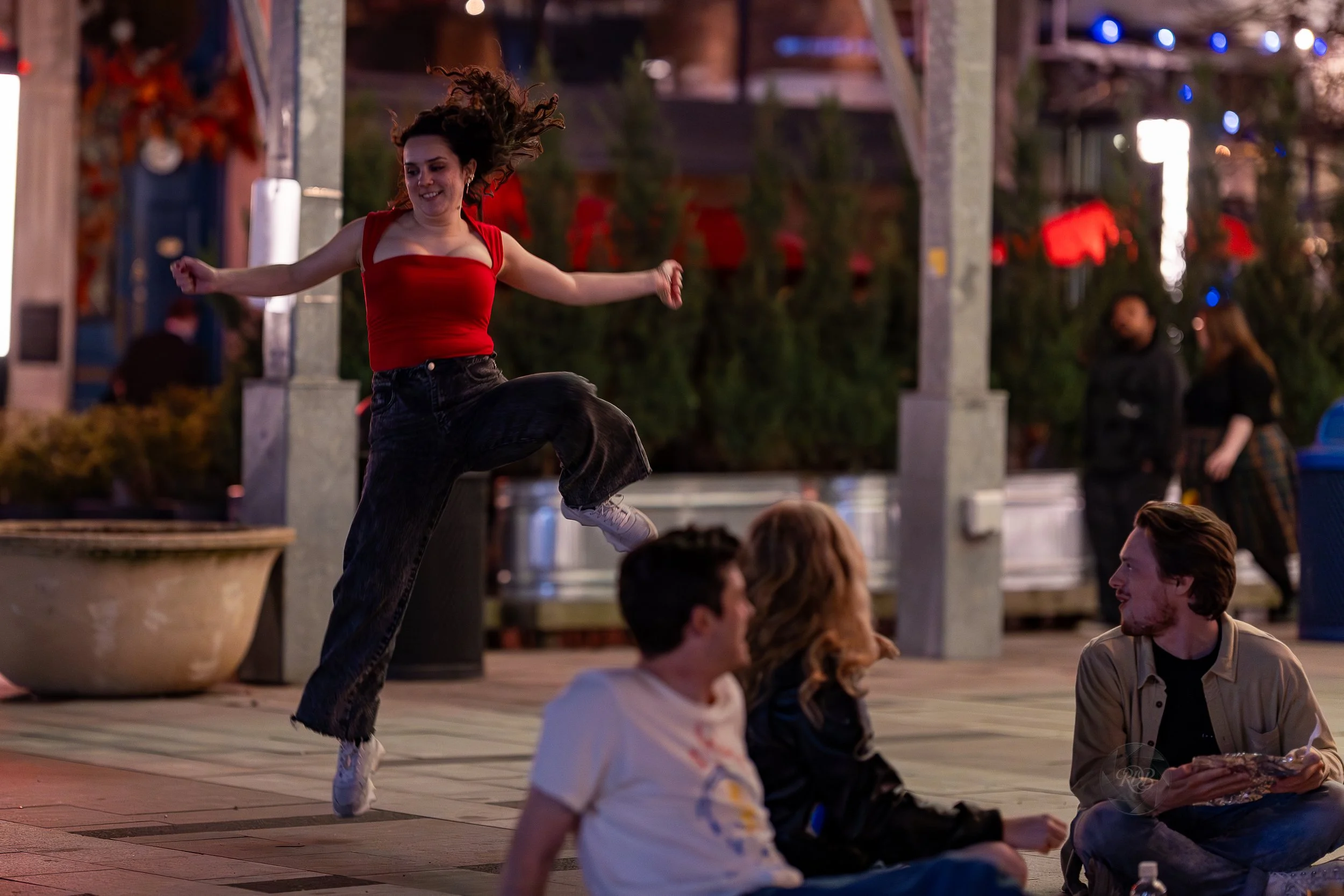 A young woman wearing a red top and black pants is jumping in the air while dancing at an outdoor evening gathering, with three people sitting on the ground nearby engaged in conversation.