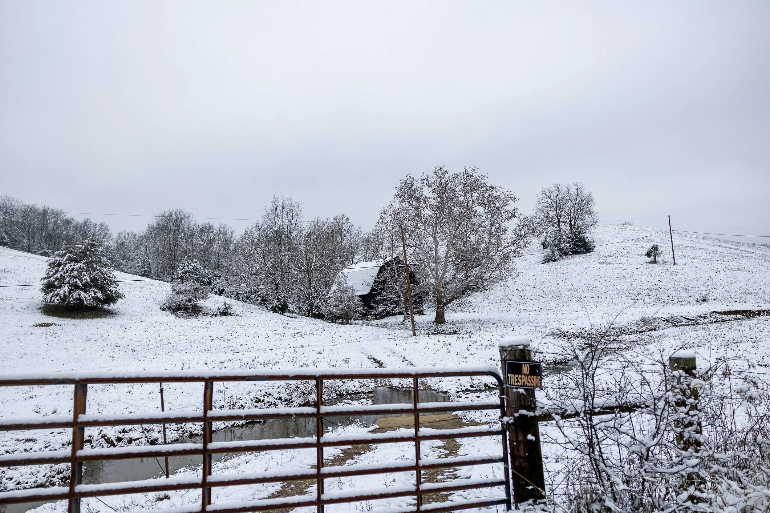 Snow-covered rural landscape with trees, a barn, a creek, and a 'No Trespassing' sign on a gate.