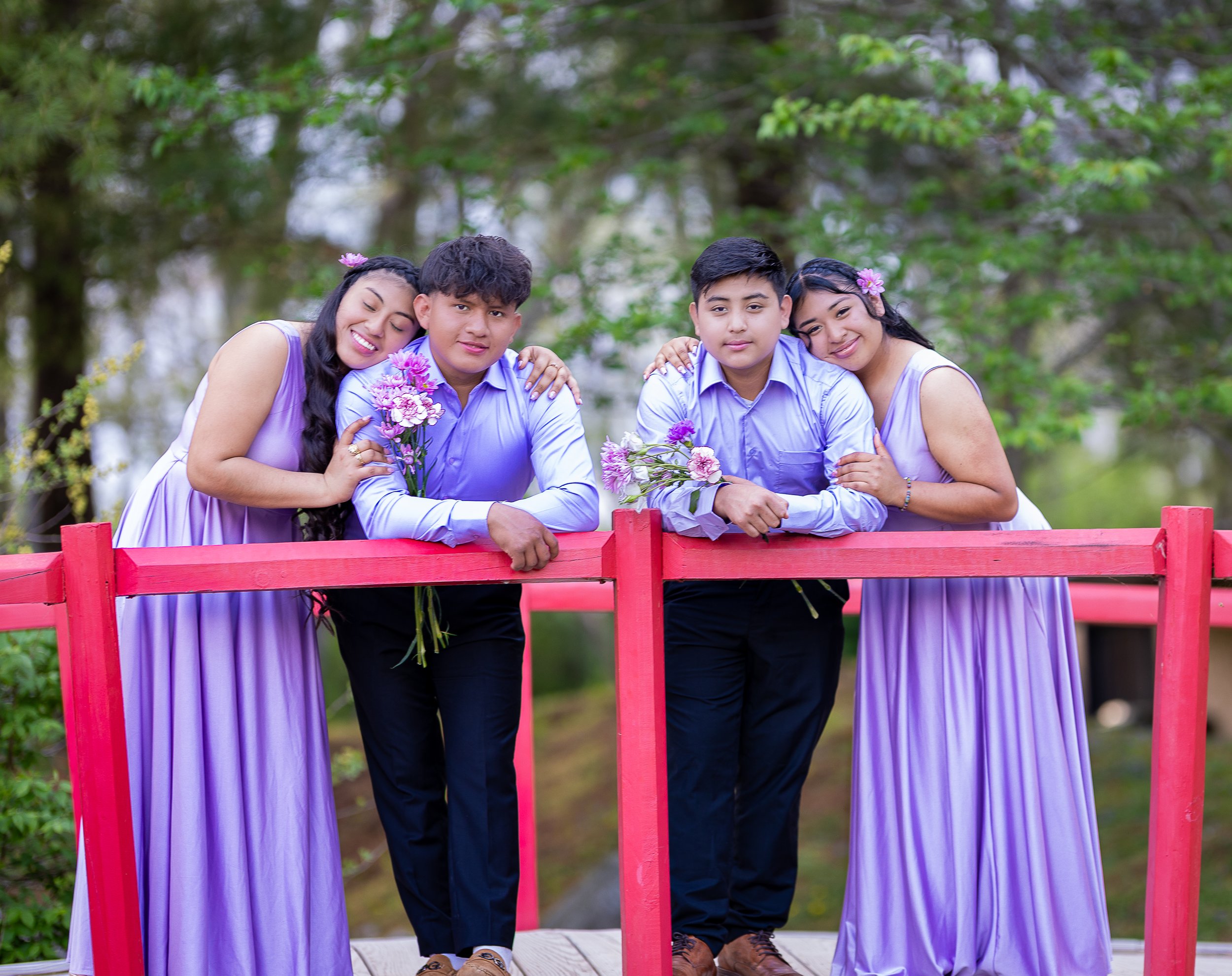 Four teenagers, two boys and two girls, standing behind a red railing outdoors, holding purple flowers and hugging each other, dressed in matching light purple shirts and lavender dresses, with trees in the background.