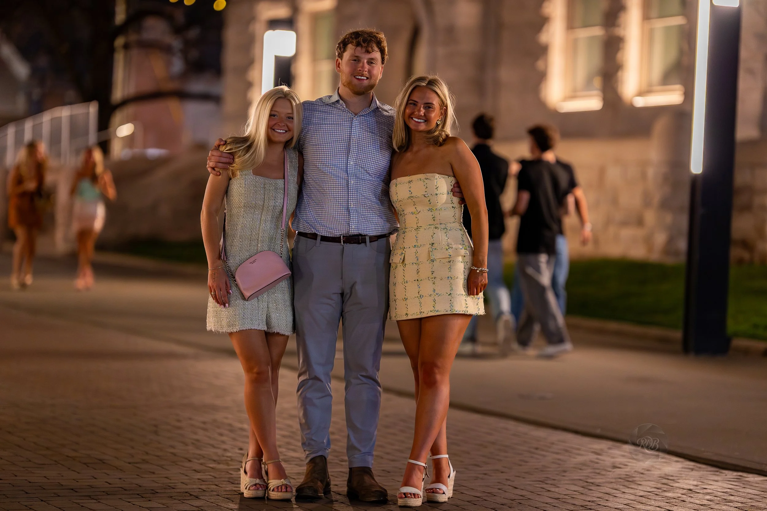 Three young adults, two women and a man, standing together on a city street at night, smiling.