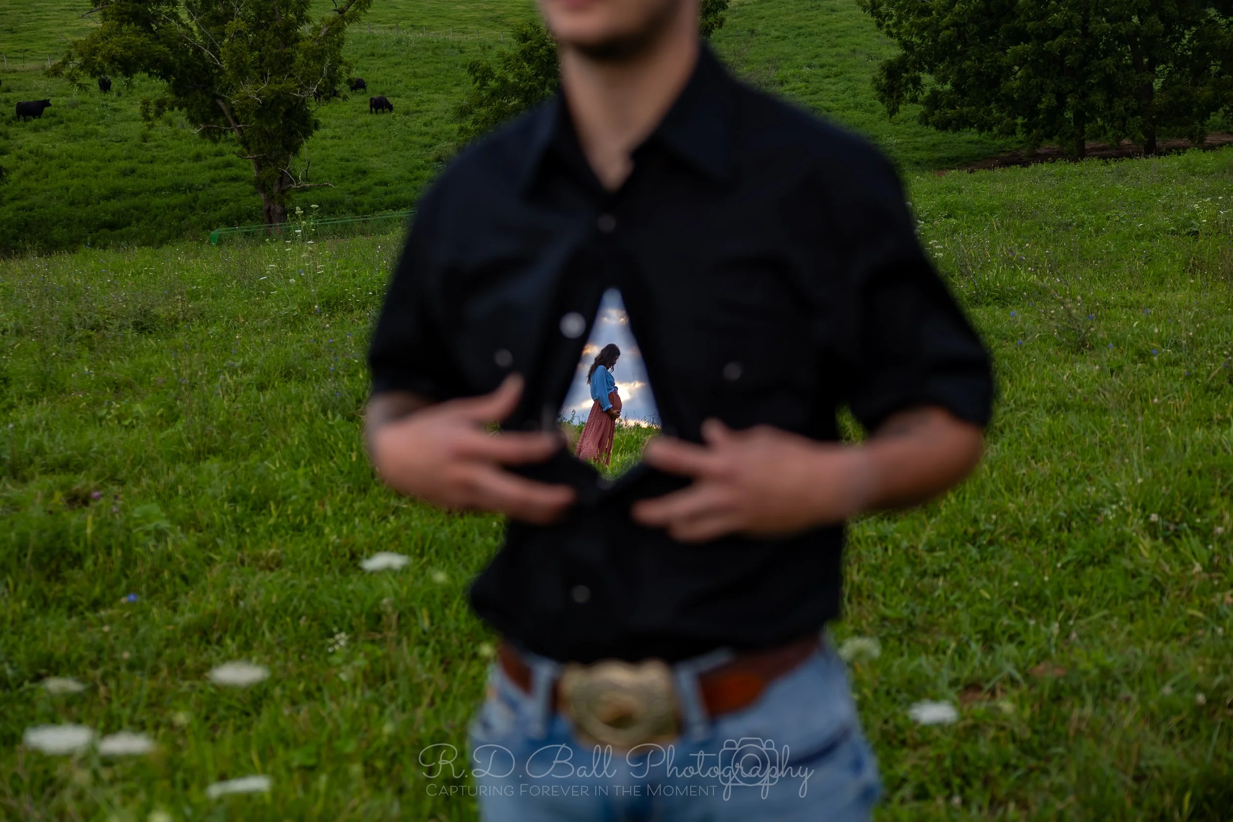The person in the foreground is holding open their black shirt to reveal a reflection of a woman in a blue shirt and brown skirt standing outdoors by a lake, with green grass, trees, and buffalo in the background.