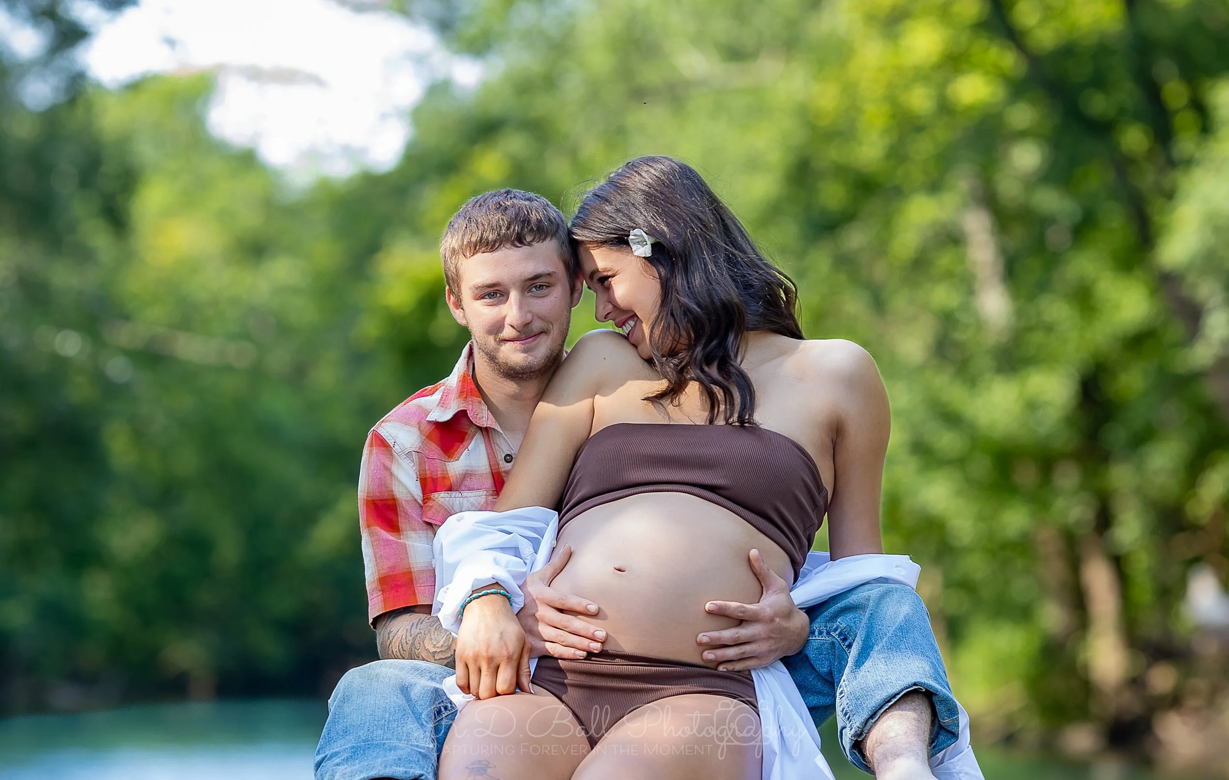 A pregnant woman and a man sitting outdoors among green trees, with the woman smiling and leaning her forehead against the man's temple. The man is holding her belly, and they both appear happy and affectionate.