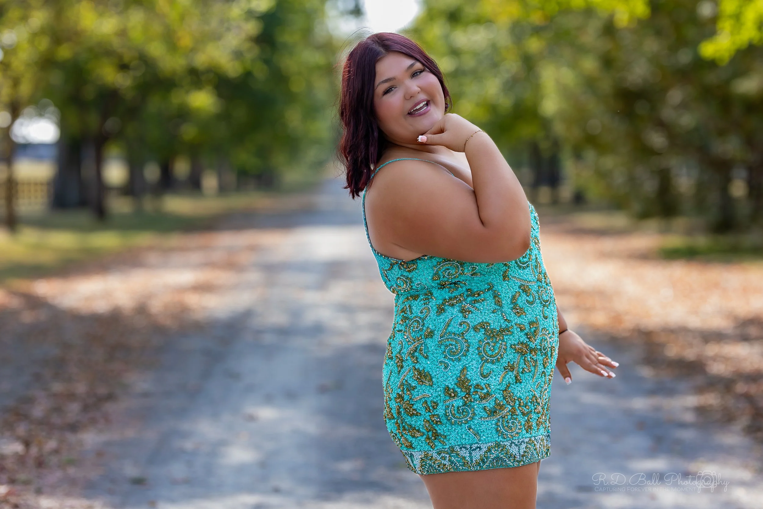 A woman in a turquoise floral dress with spaghetti straps, smiling and posing outdoors on a tree-lined path.