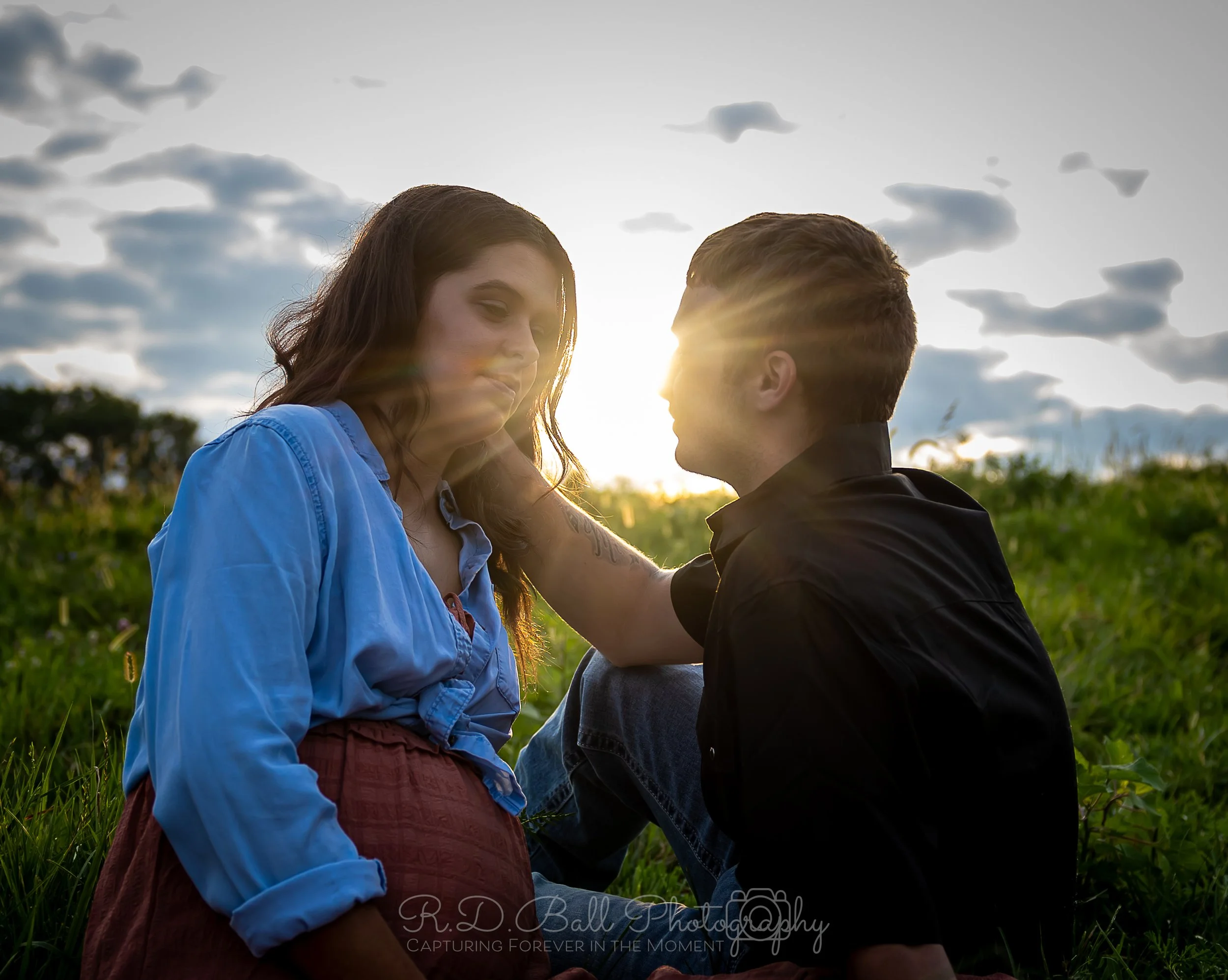 A loving couple sitting on grass in a field at sunset, gazing at each other, with the sun shining behind them and clouds in the sky.