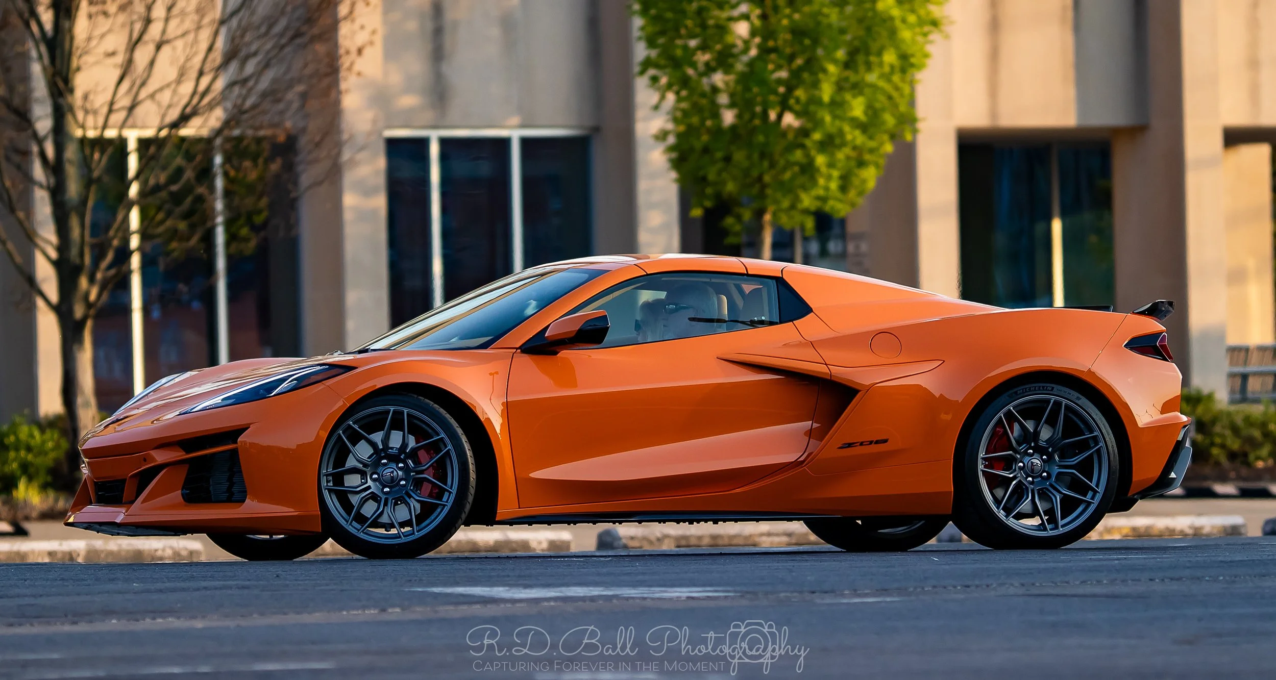 Orange sports car parked on a city street with modern buildings and trees in the background.