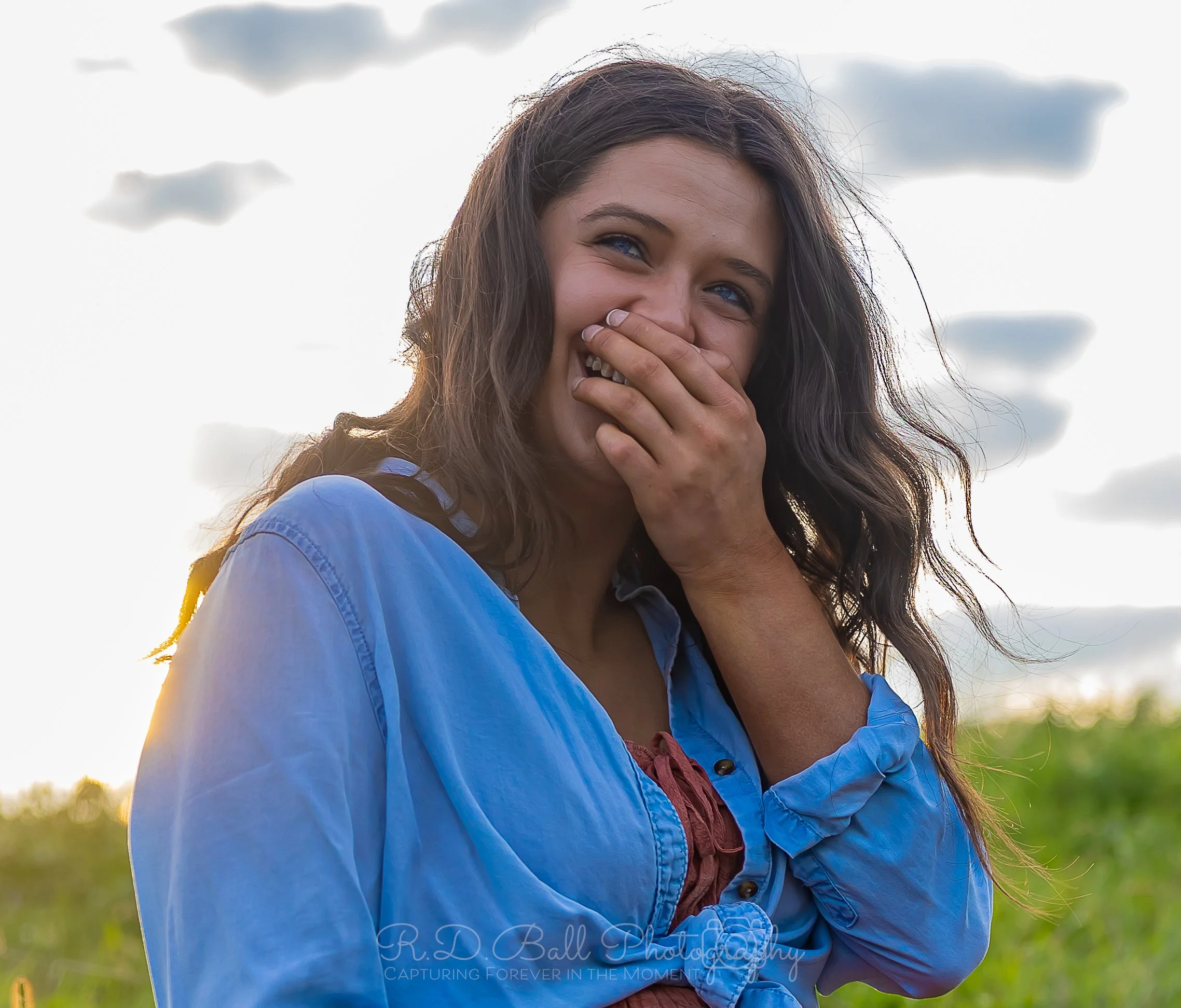 A woman with long wavy brown hair smiling and covering her mouth with her hand, wearing a blue shirt, outdoors with a cloudy sky in the background.