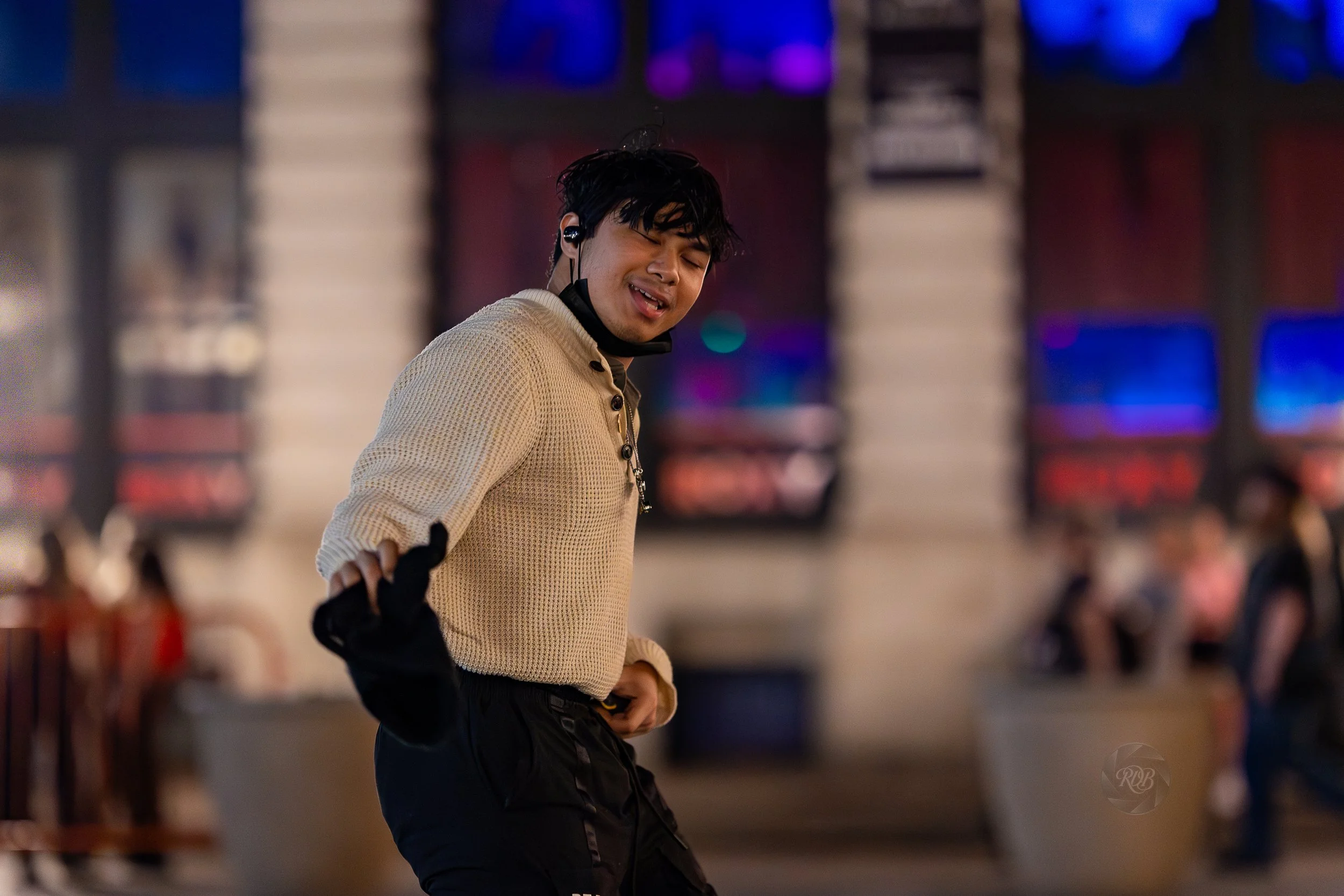 A man with black hair wearing a beige sweater and black gloves stands in a lively indoor space with colorful lights and blurred background.