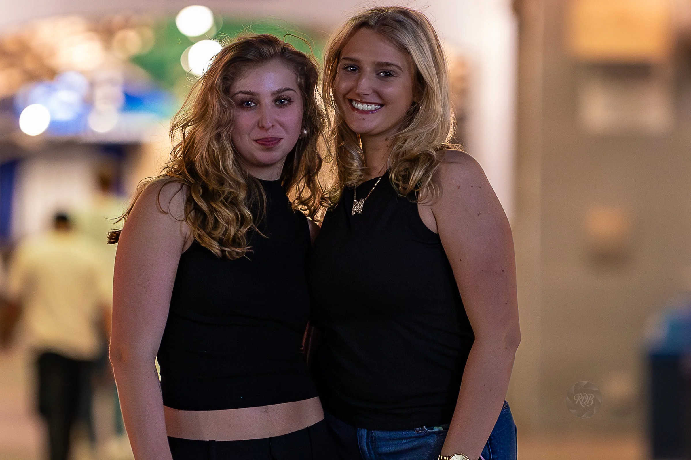 Two young women with blond and light brown hair smiling and standing close together in a lively setting at night, wearing black sleeveless tops.