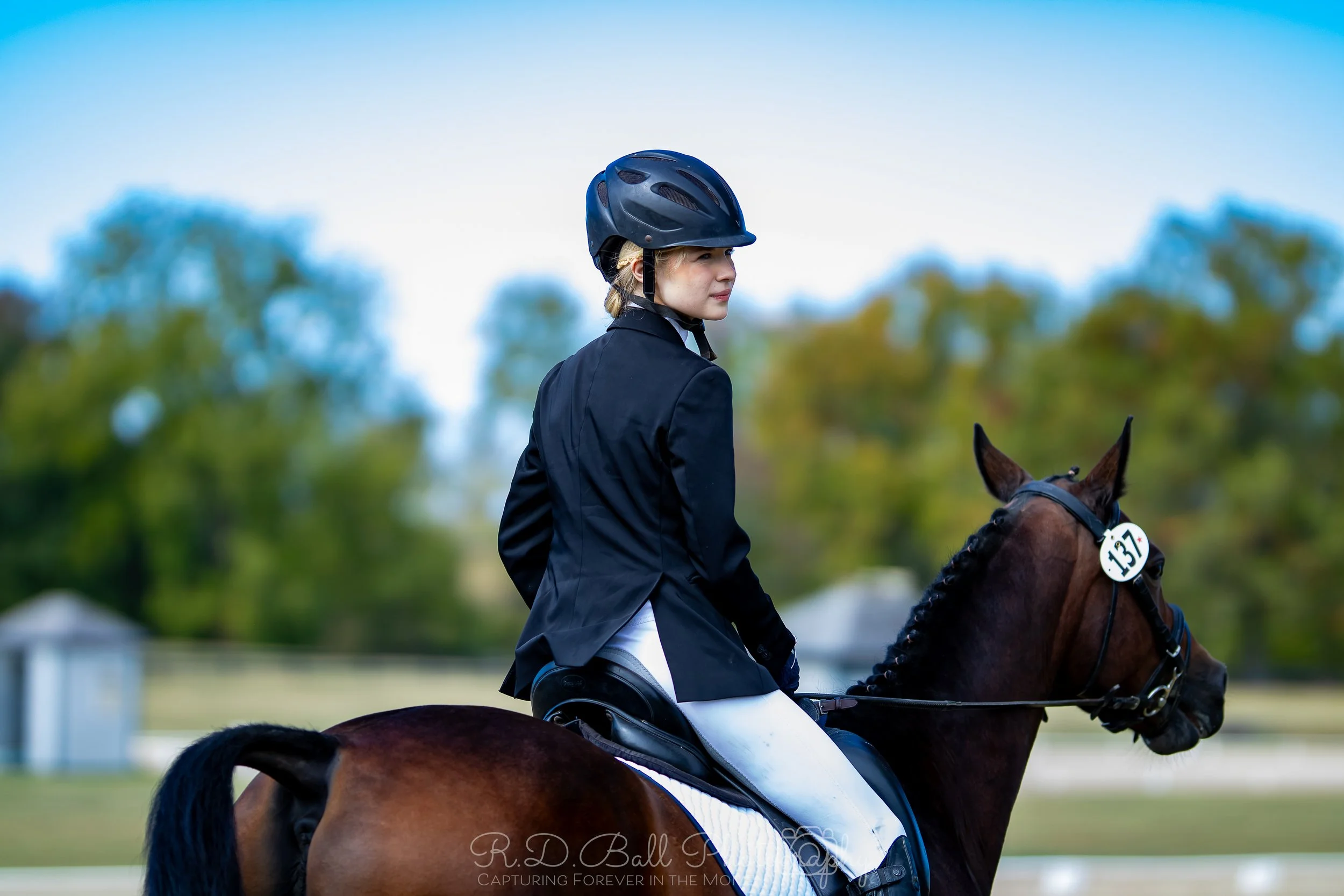 Young woman in equestrian attire riding a horse during a show, with a helmet, black jacket, and white riding pants.