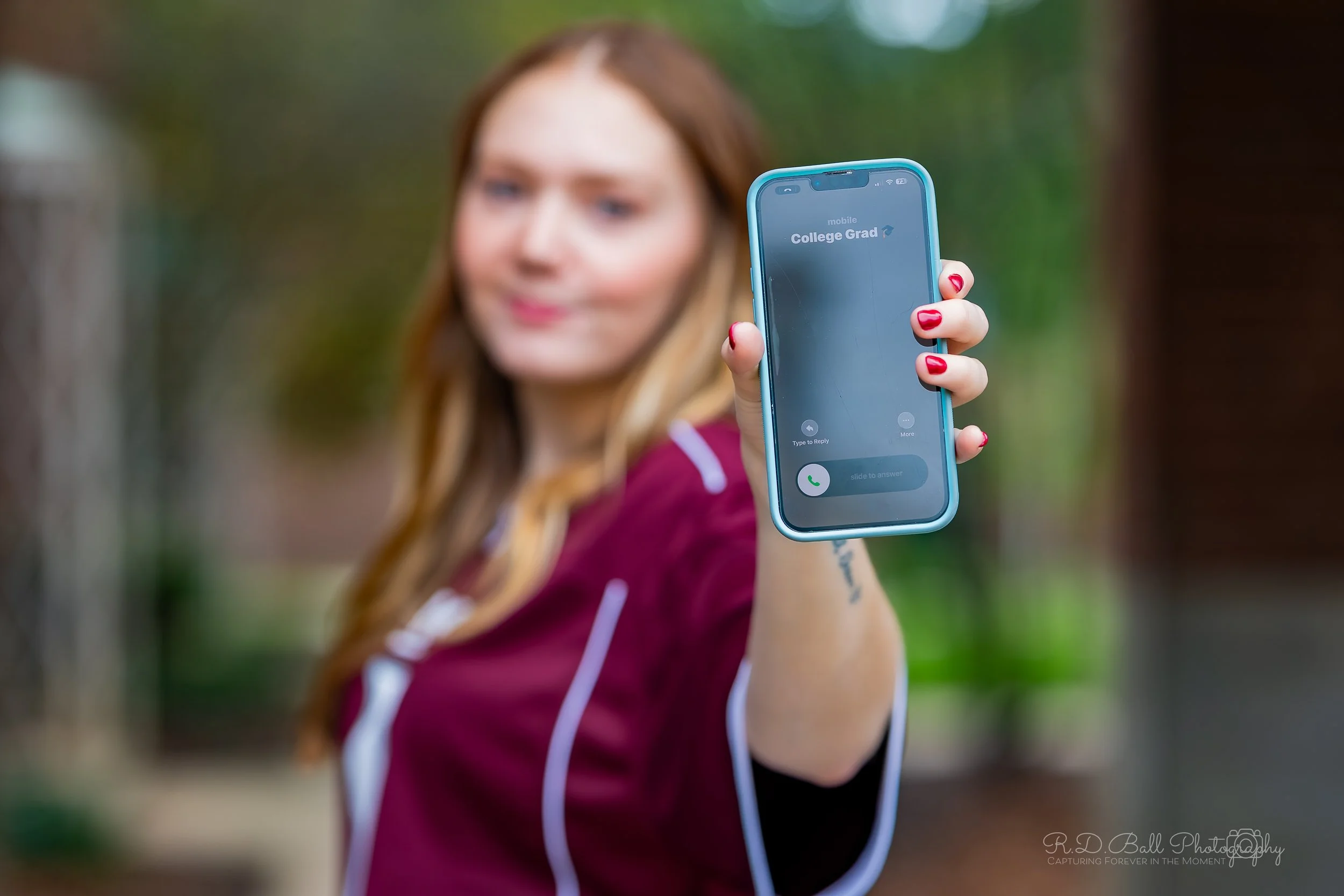 A woman with red hair holding a blue smartphone displaying an incoming call from 'College Grad' outside in a park.