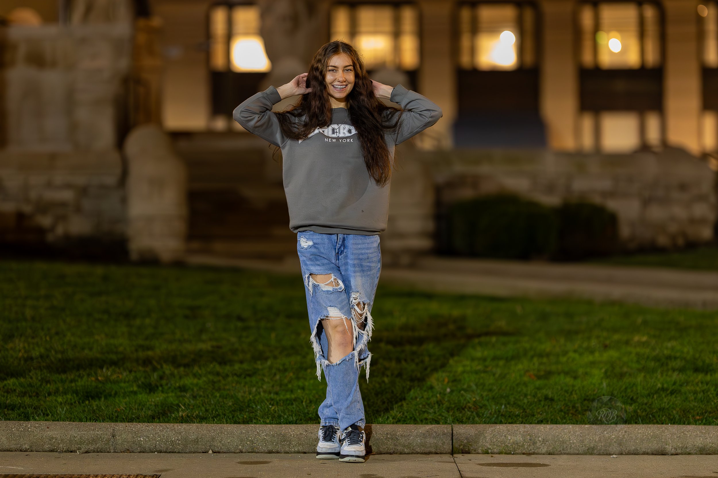 Young woman with long brown hair, smiling, standing outdoors at night on a sidewalk with a grassy area, wearing a gray sweatshirt and ripped jeans.