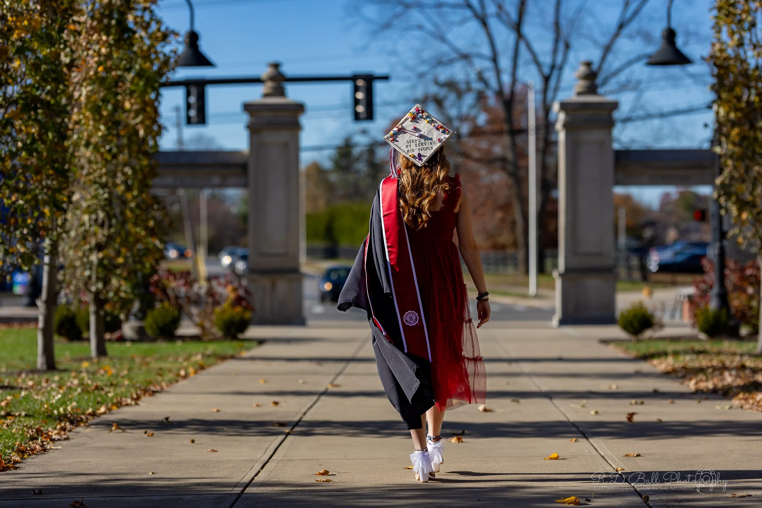A woman wearing a graduation gown and cap walking on a sidewalk, with trees and an archway in the background on a sunny day.