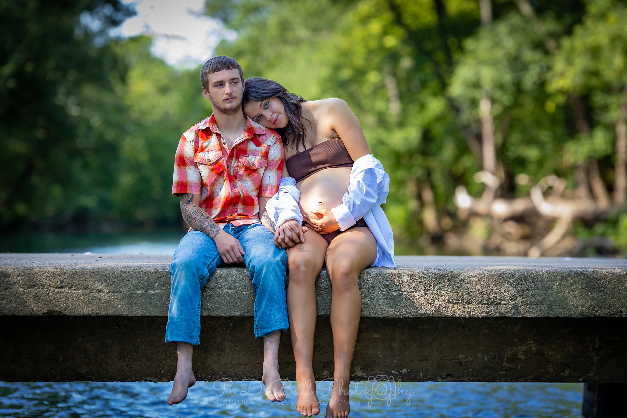 A young couple sitting on a concrete dock by the water, with the woman pregnant. They are holding hands, with the woman resting her head on the man's shoulder, surrounded by green trees.