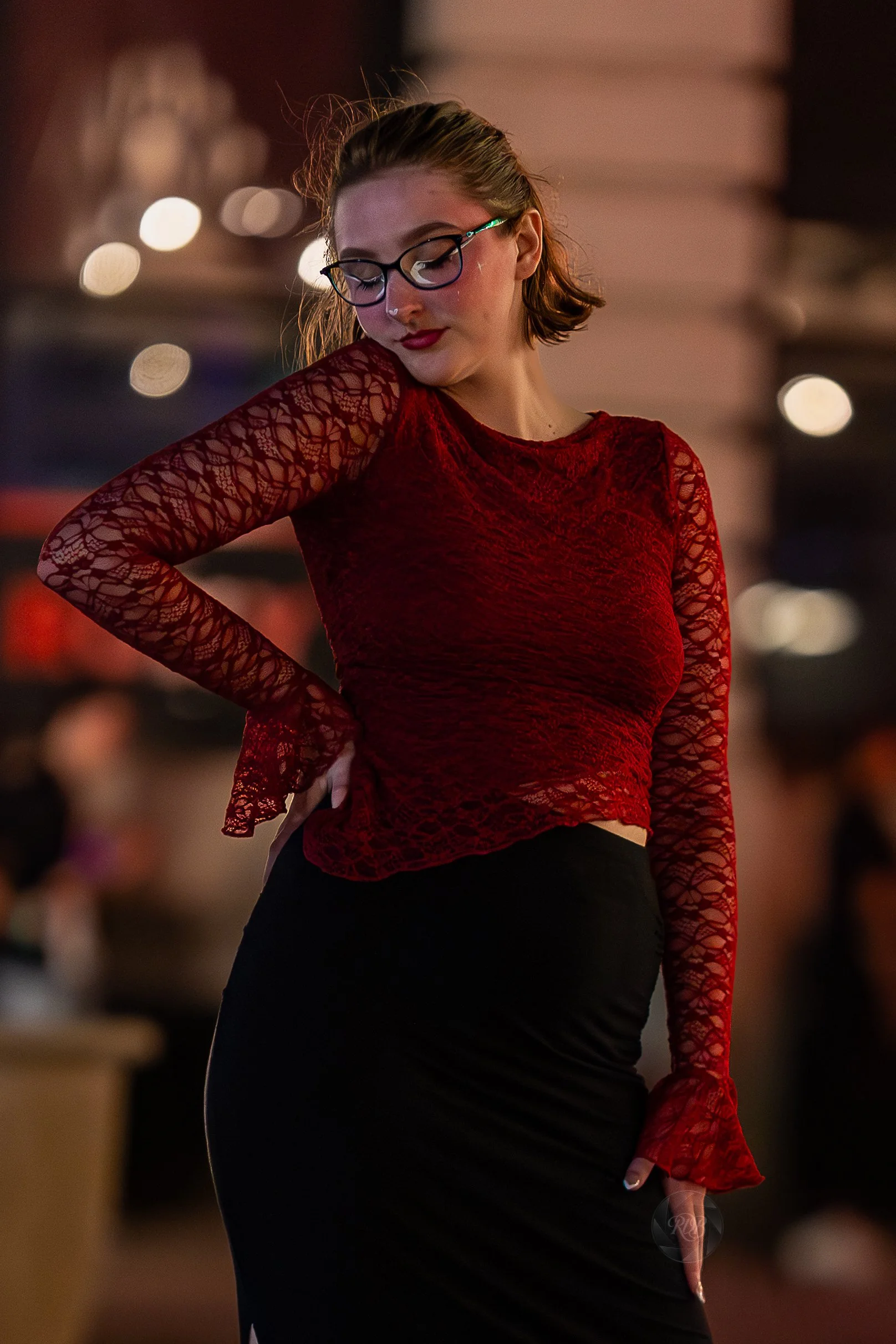 A woman in a red lace top and black skirt, wearing glasses, looking down with her hand on her hip in a dimly lit indoor setting.