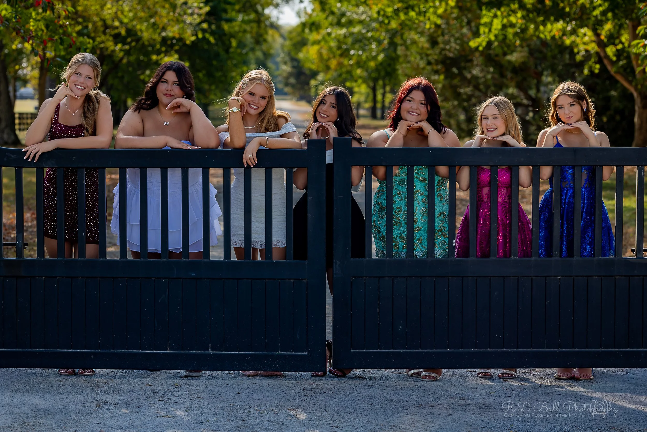Seven young women in colorful dresses stand behind a black gate on a sunny day in a park with green trees in the background.
