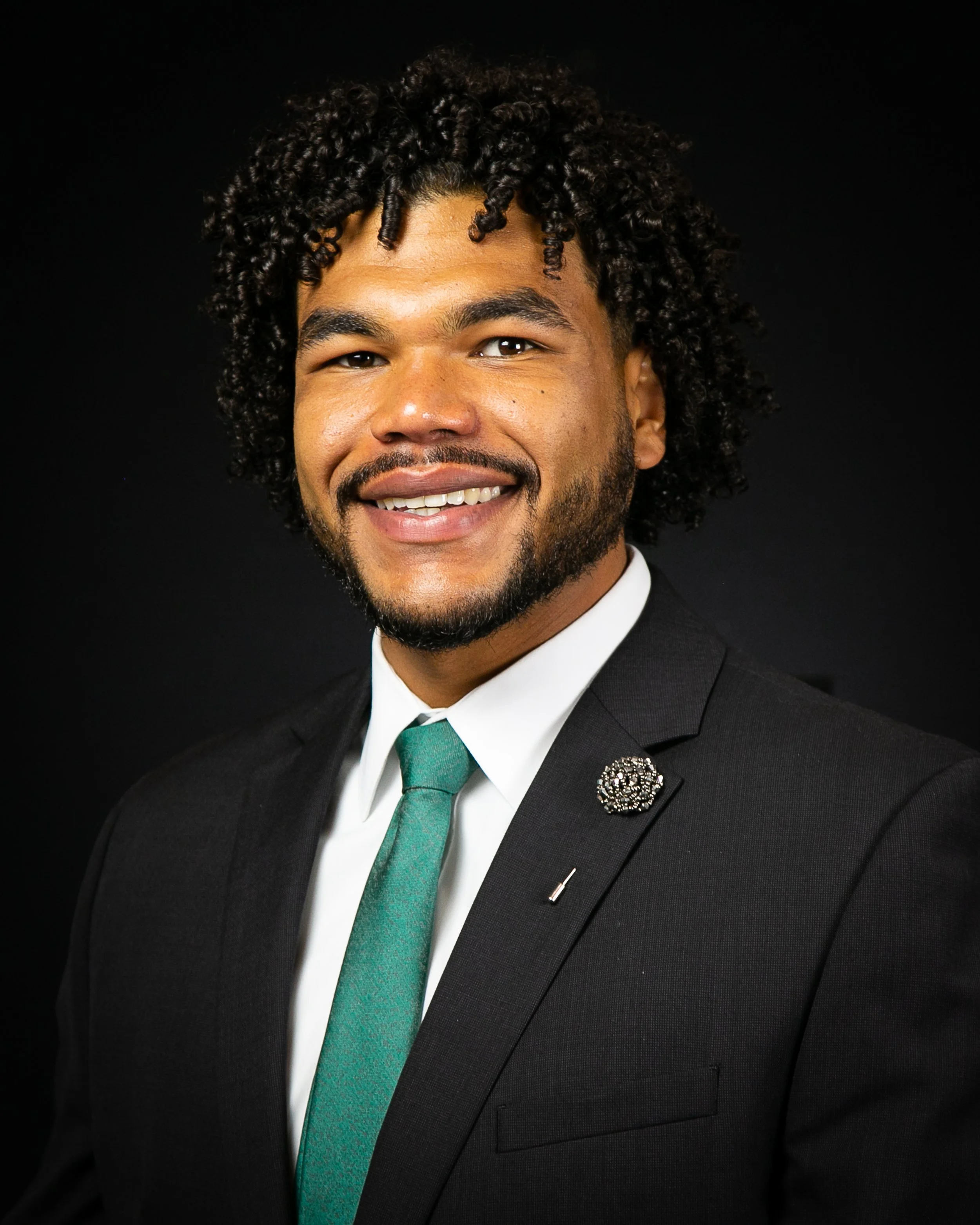 A smiling man in formal attire wearing a black suit, white shirt, and teal tie, with a decorative pin on his lapel, against a black background.