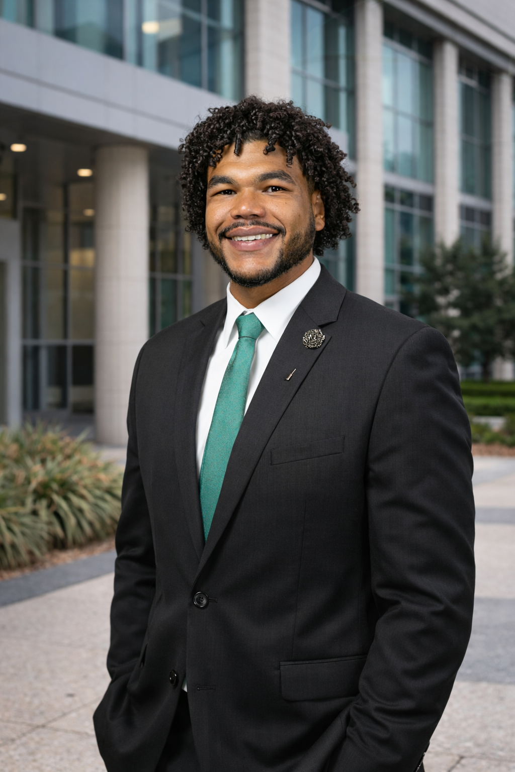 A smiling man in a suit with a teal tie, standing outside a modern office building.