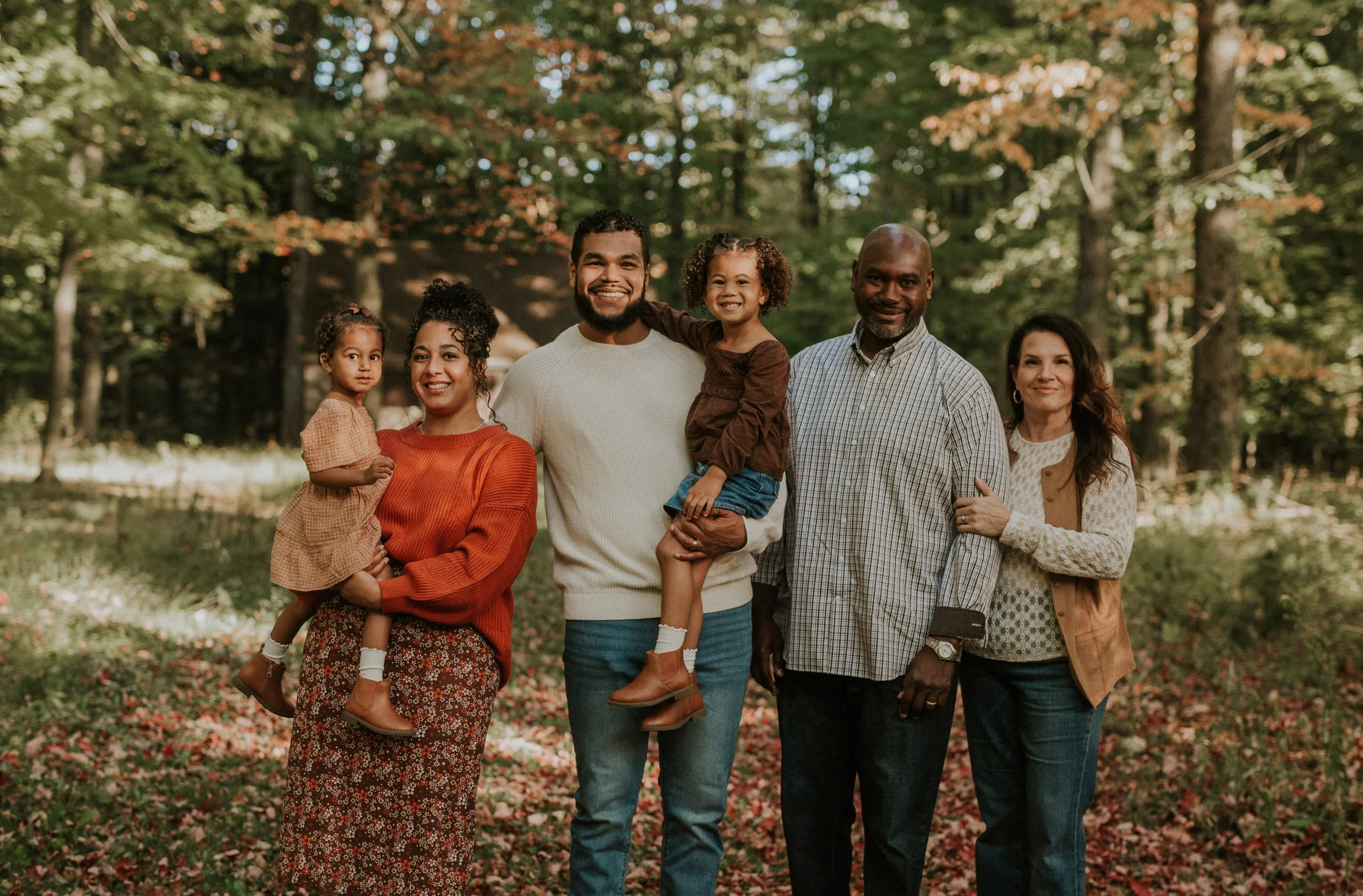 A multi-generational African American family of six smiling and posing outdoors in a wooded area during autumn.