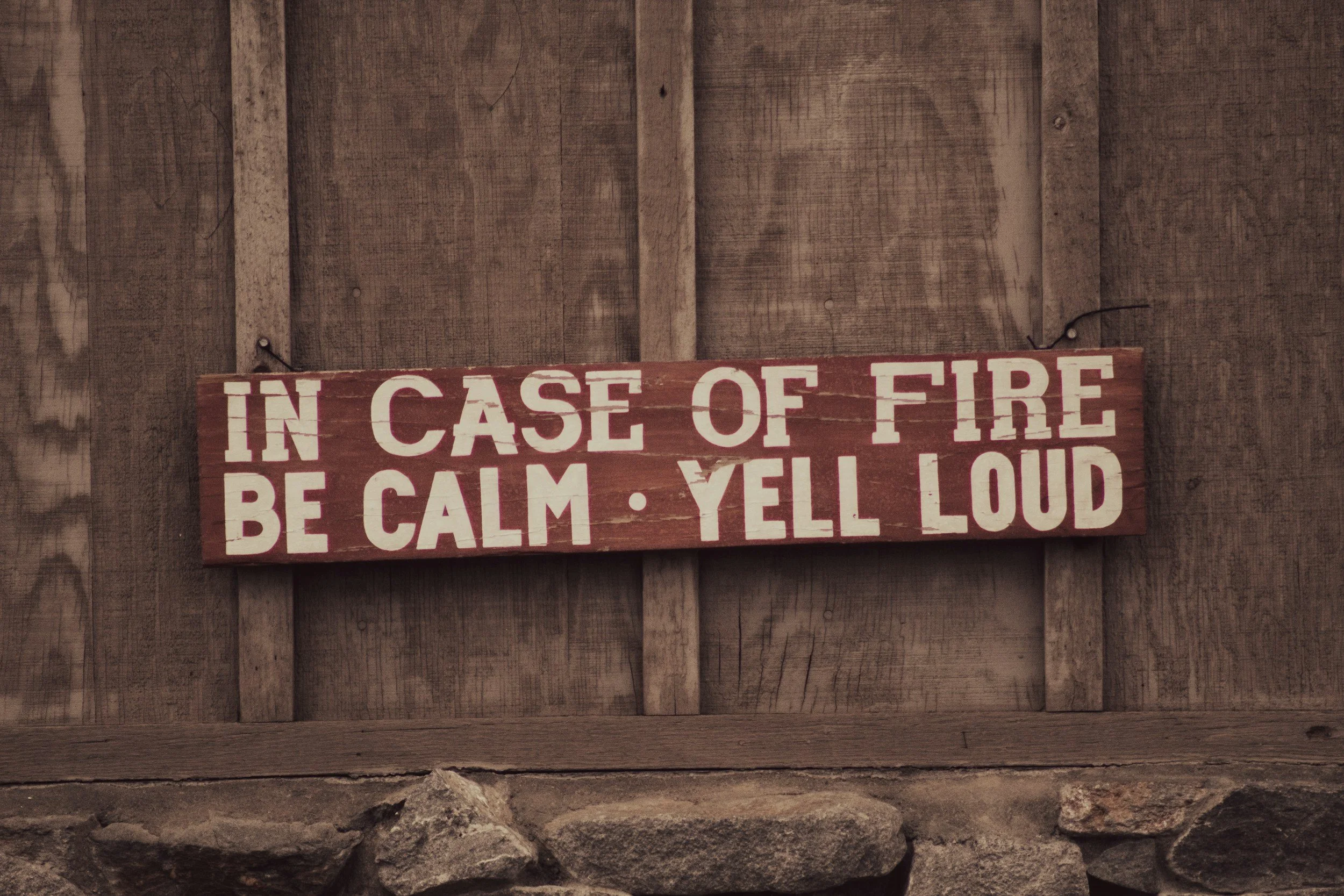 A wooden sign with red background and white text saying "In case of fire be calm yell loud" hangs on a rustic wooden wall.
