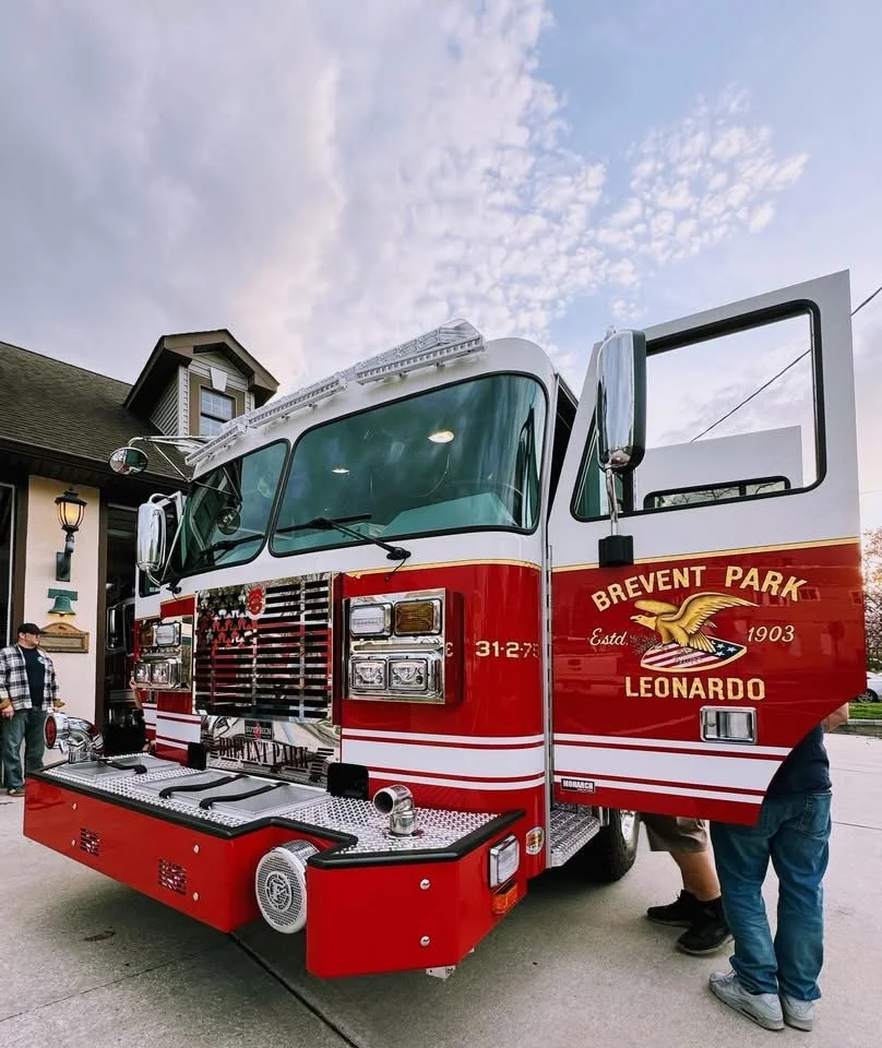 A red fire truck from Brevent Park Leonardo fire department parked outdoors with two people inspecting it. The fire truck has a white top and front, with the logo and text on the side. The background shows a house and a cloudy sky.