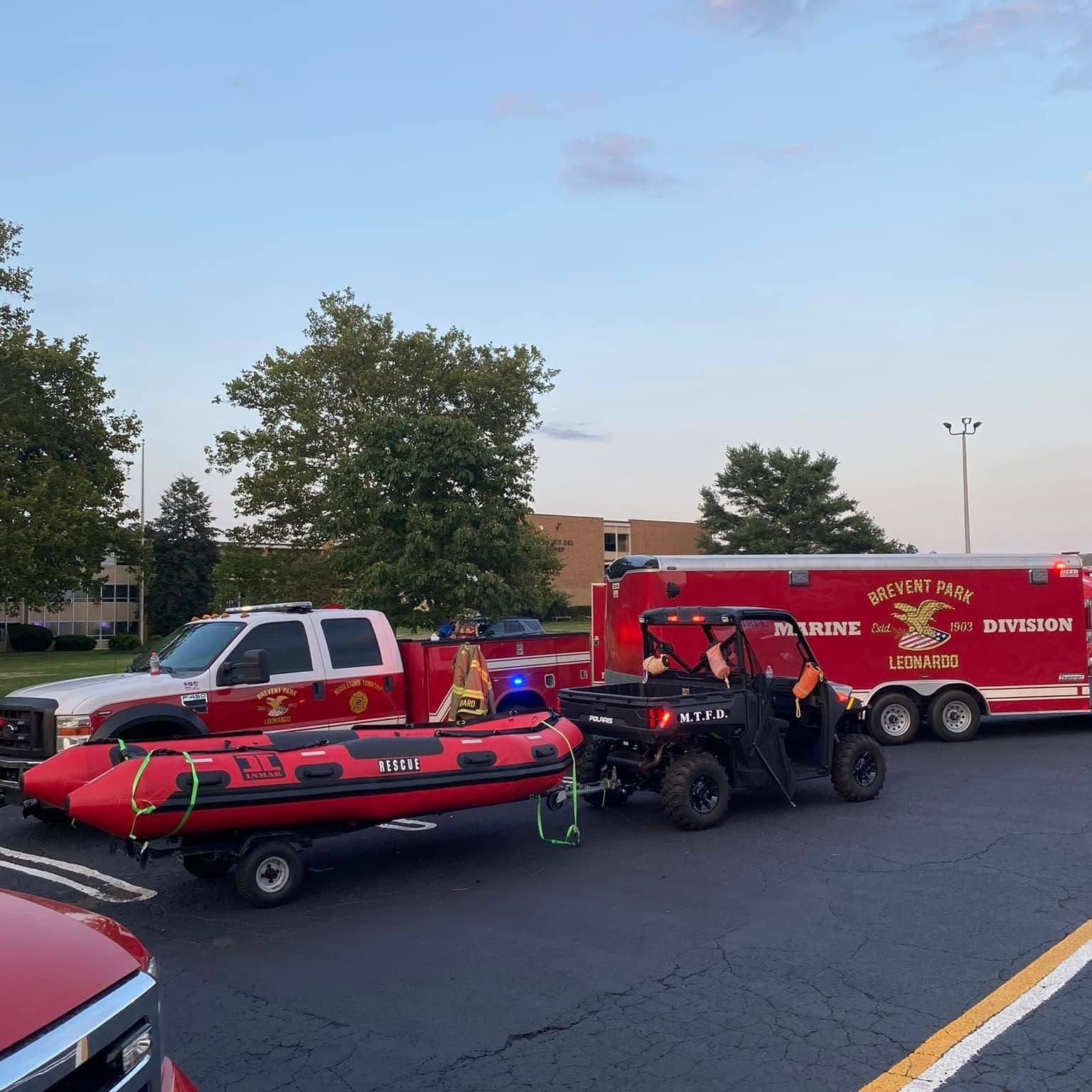 A fire rescue vehicle, a fire department utility vehicle, a rescue boat on a trailer, and a small off-road utility vehicle are parked in a parking lot with trees and a building in the background.