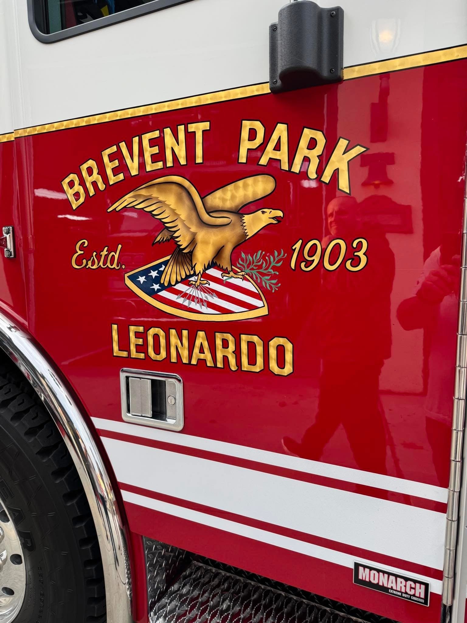 Close-up of red fire truck door with logo 'Brewent Park, Estd. 1903, Leonardo' featuring an eagle holding a branch and shield with American flag design, and a Monarch brand badge.