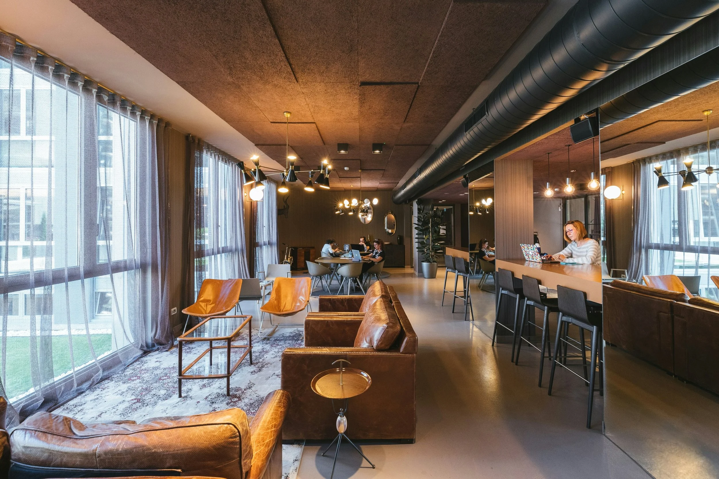 A modern, stylish lounge or coworking space with leather armchairs, bar stools at a counter, and large windows with sheer curtains. Several people are working on laptops, and the space is illuminated by contemporary hanging lights.