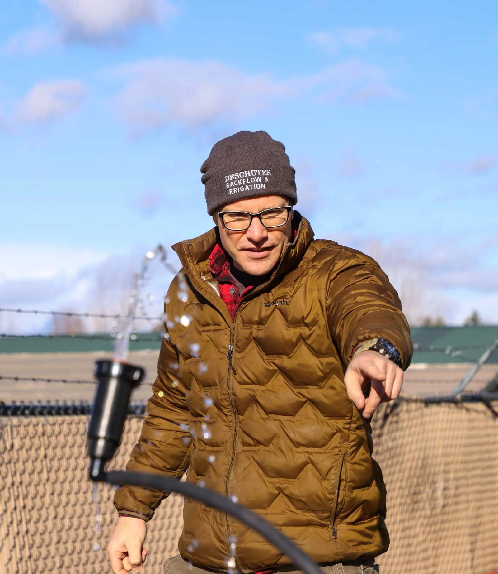 A man wearing glasses, a brown jacket, a gray beanie, and a red plaid shirt underneath, points at the camera outside on a sunny day with a chain-link fence and blue sky in the background.