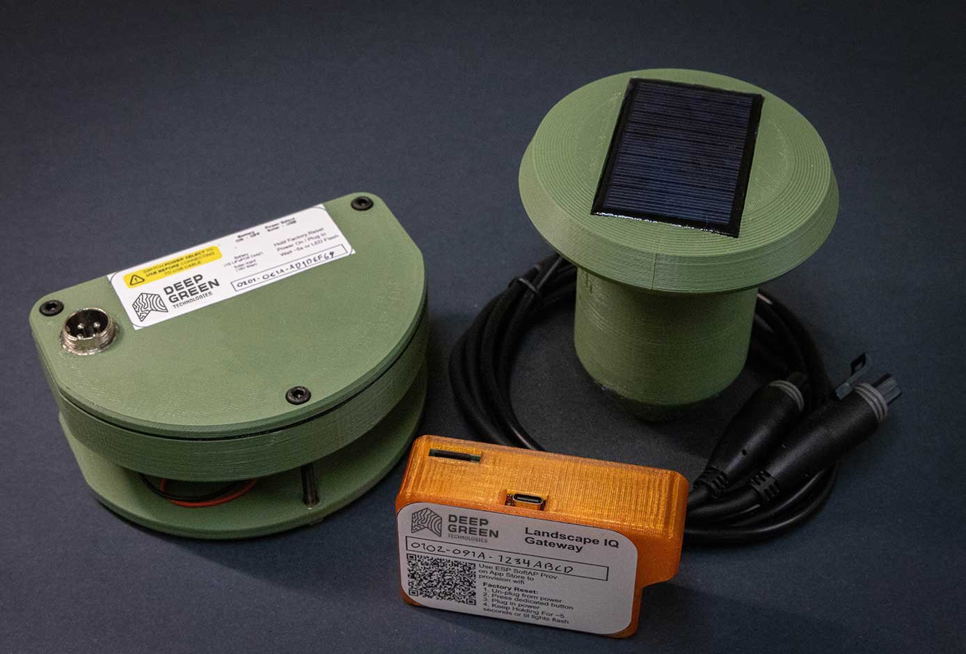Three green electronic devices with black cables, one has a solar panel on top, and the smallest is orange labeled 'Landscape IQ Gateway' from Deep Green Technologies.