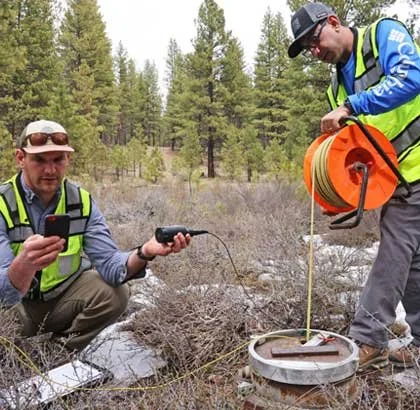 Two men in safety vests using equipment outdoors in a forested area with trees, one holding a device and the other handling a large orange reel connected to a river or wetland ground station.
