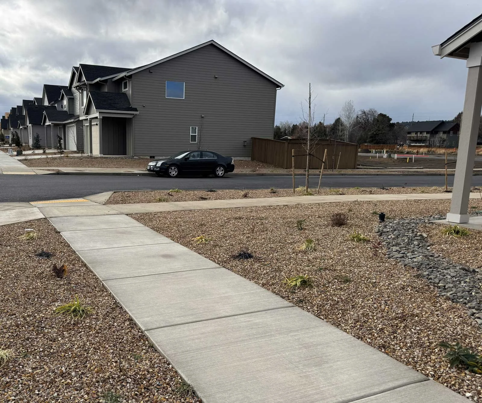 View of a residential neighborhood with modern houses, a black car parked on the street, and a partially landscaped yard with small plants and gravel rocks, under a cloudy sky.