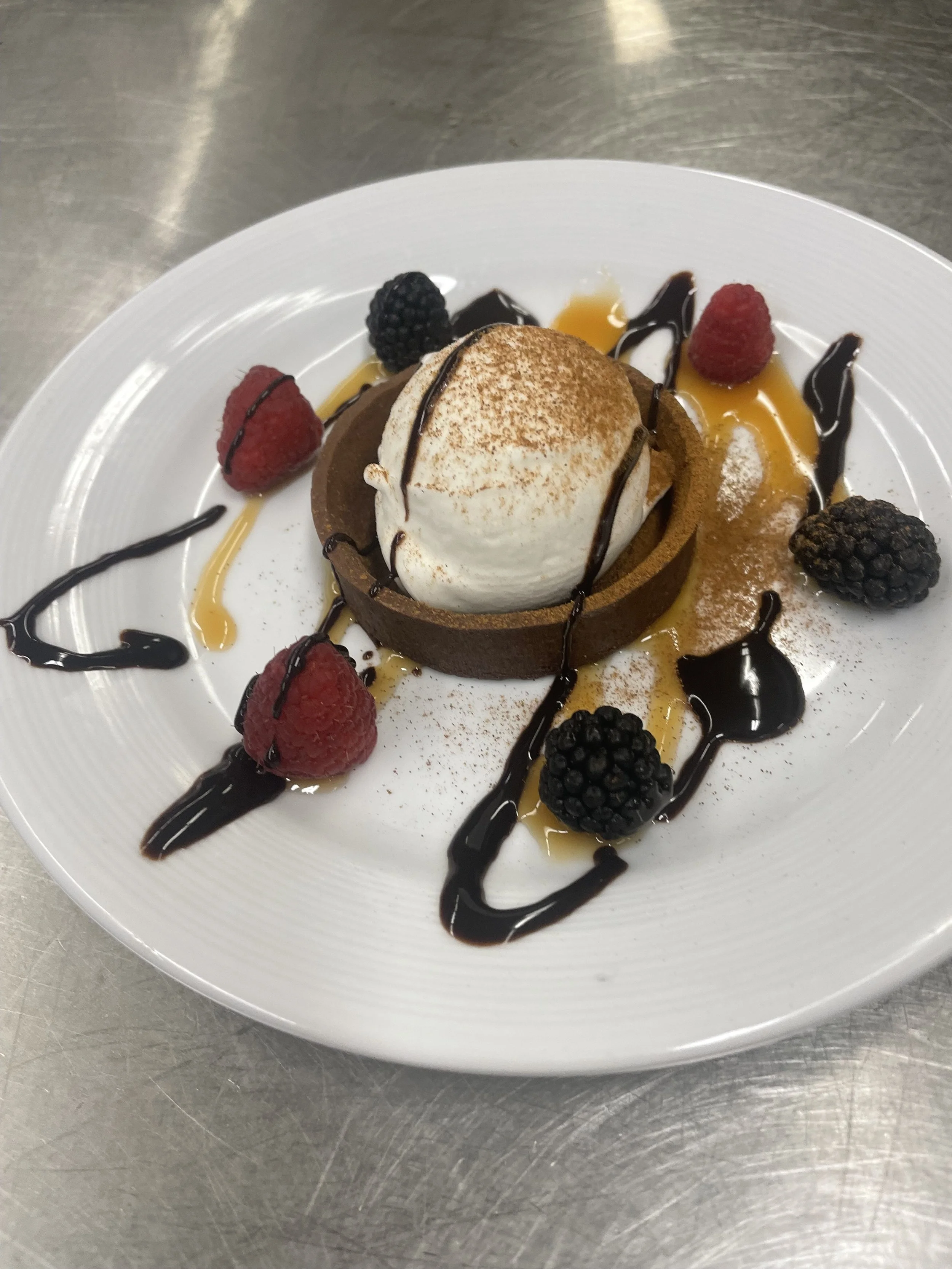 Dessert with a chocolate tart topped with a scoop of vanilla ice cream, garnished with fresh raspberries and blackberries, drizzled with chocolate and caramel sauces on a white plate.