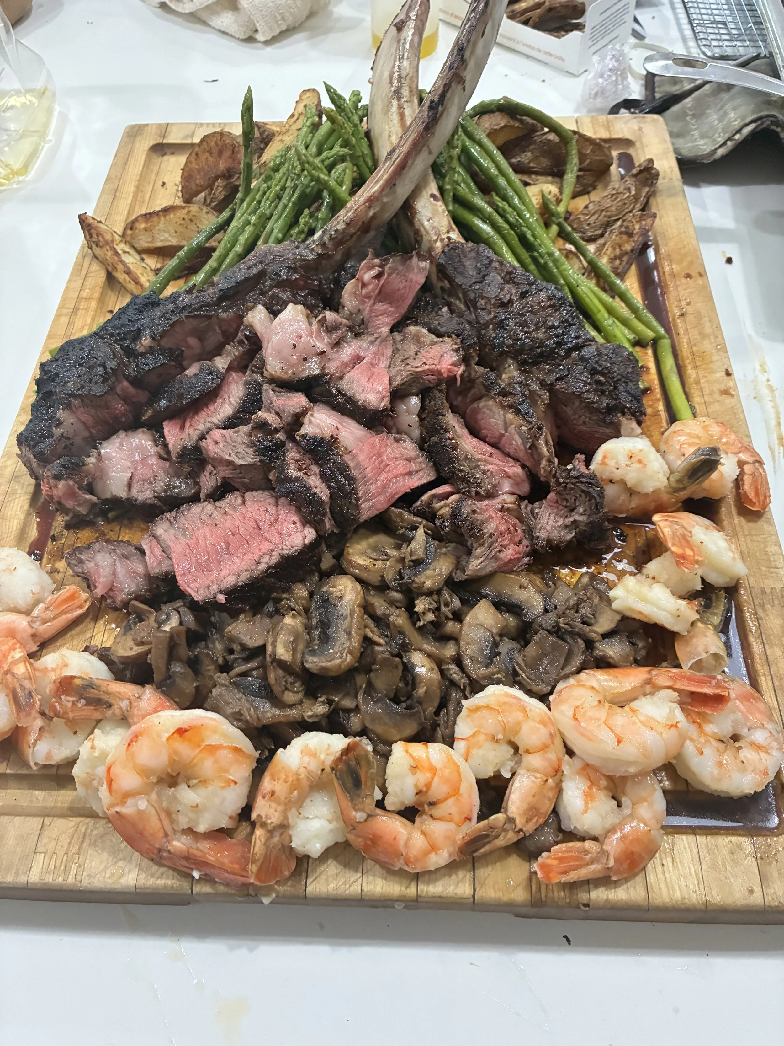A wooden board topped with sliced grilled steak, cooked shrimp, sautéed mushrooms, green beans, potato wedges, and lamb chops.