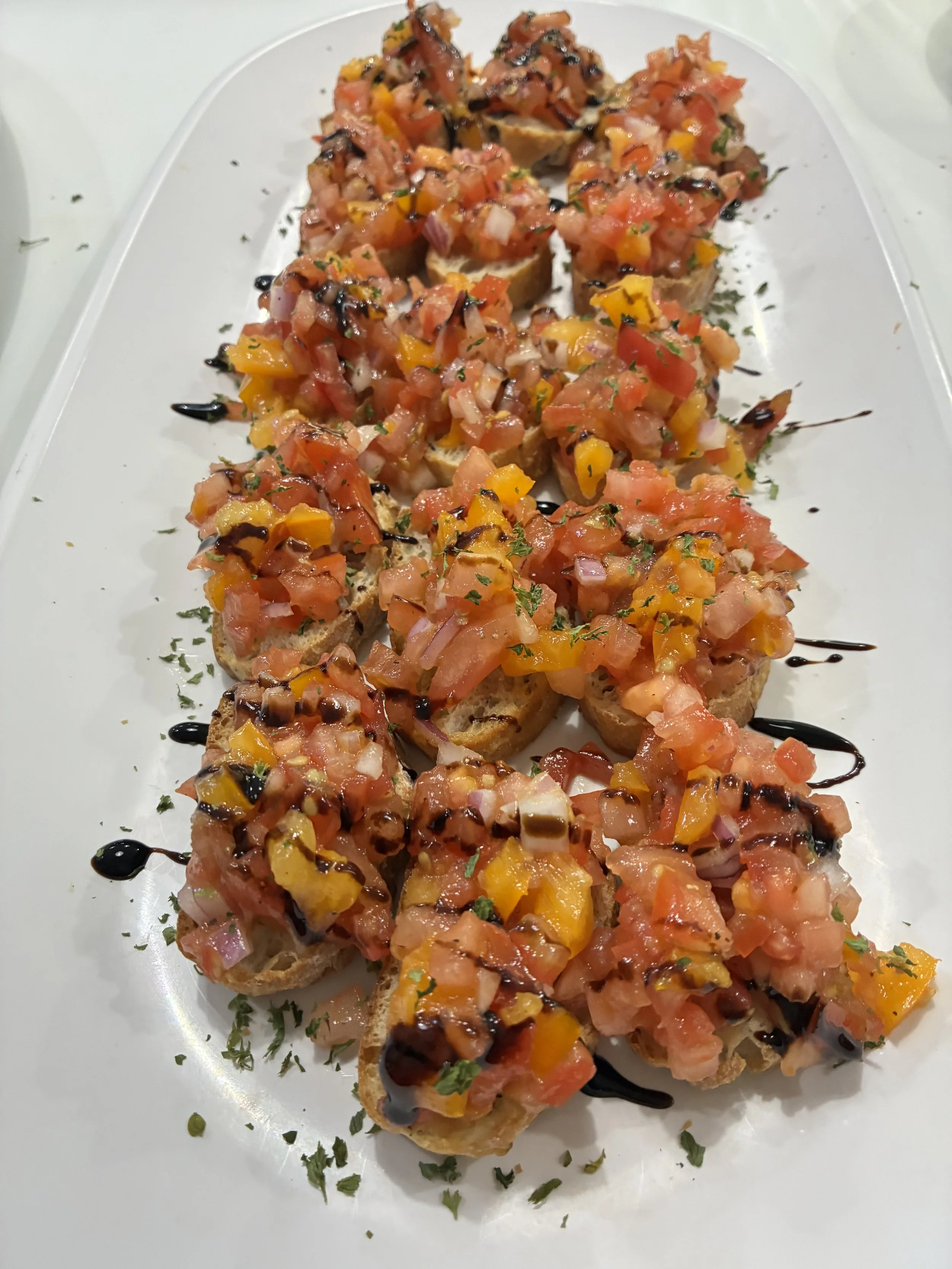 Crostini topped with a colorful mixture of diced tomatoes, yellow and red peppers, chopped herbs, drizzled with balsamic glaze, on a white serving plate.