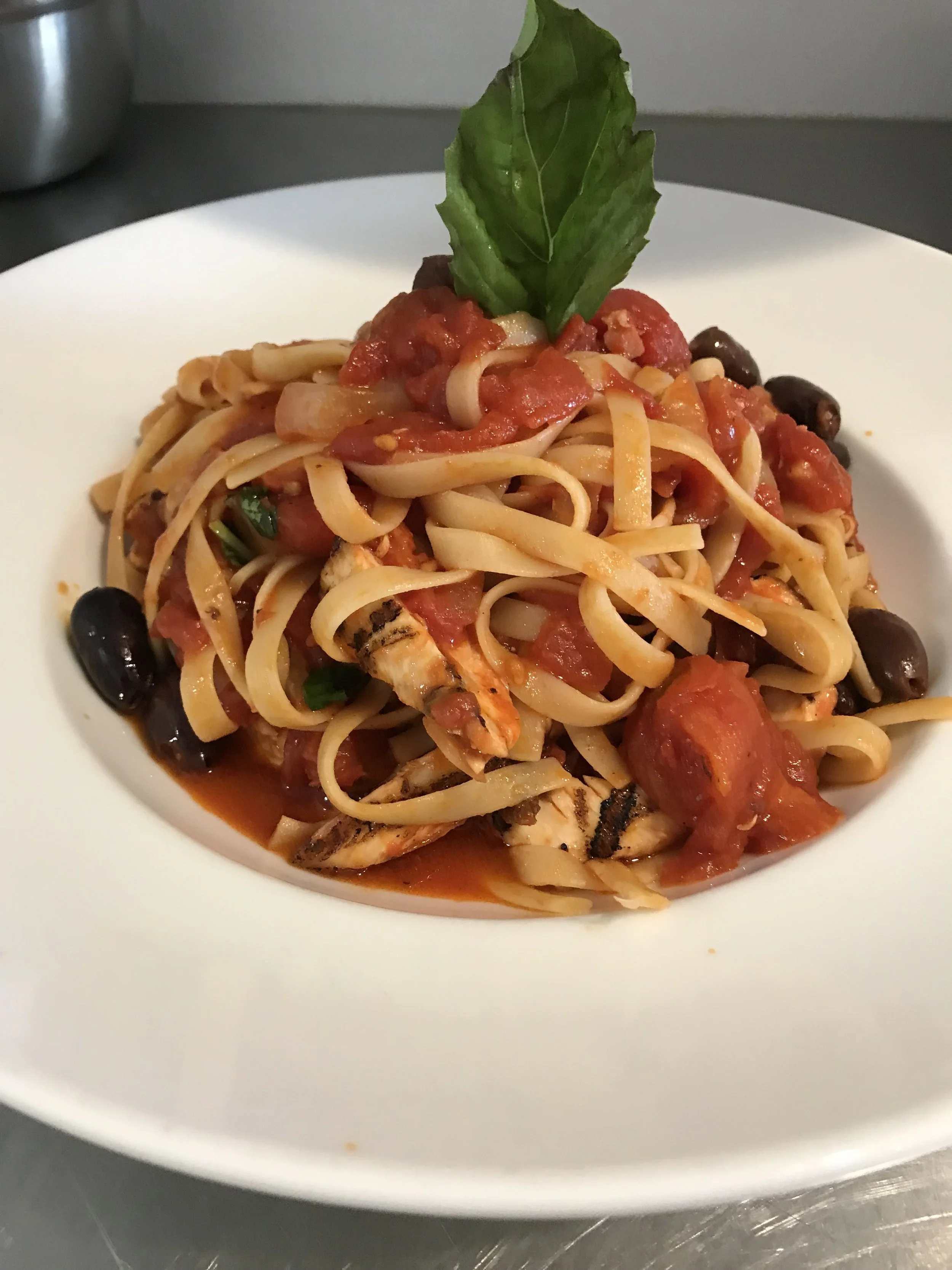 Plate of pasta with tomato sauce, black olives, grilled chicken, and a basil leaf garnish.