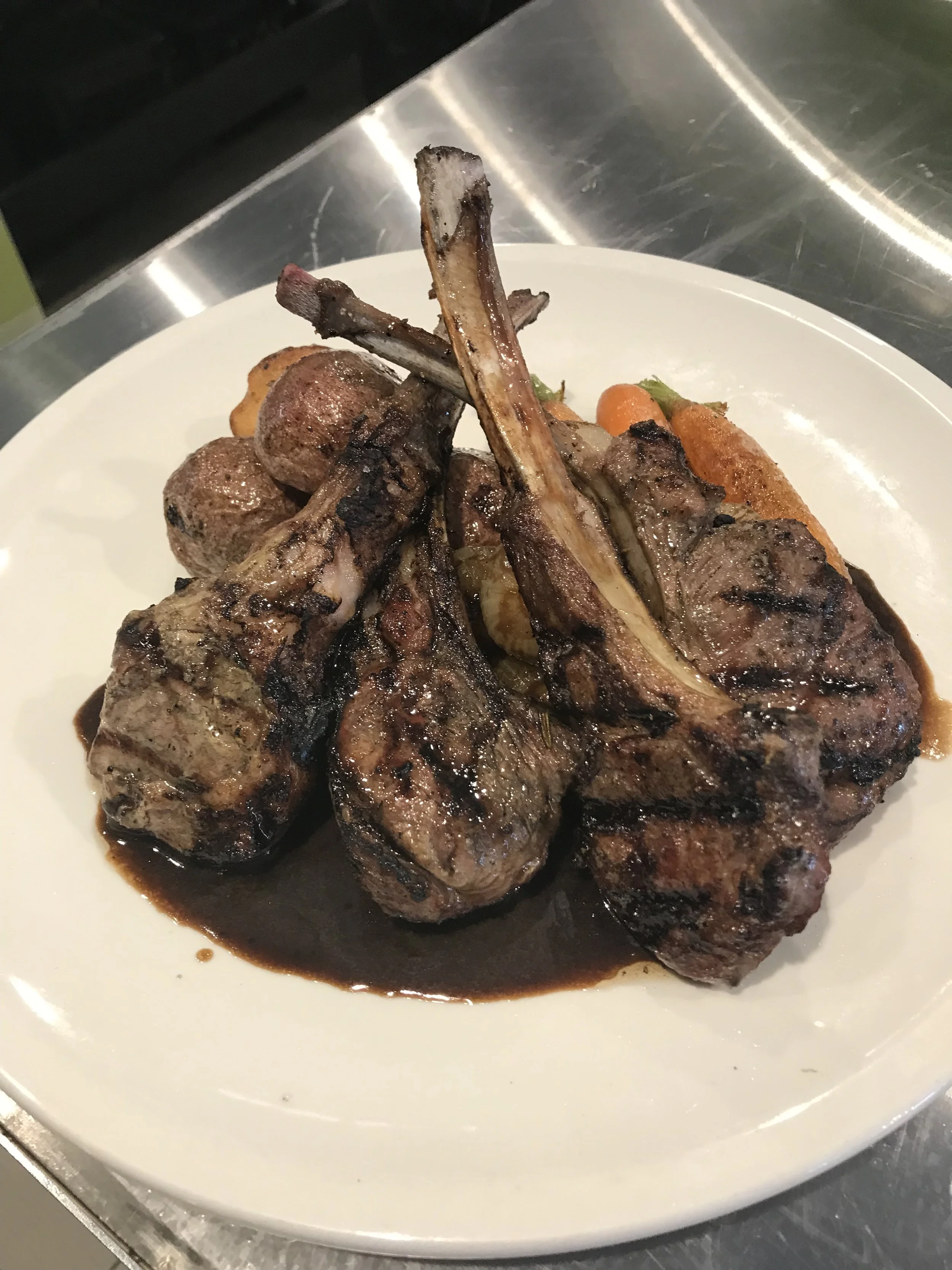 A plate of grilled lamb chops with a dark sauce, roasted carrots, and potatoes.