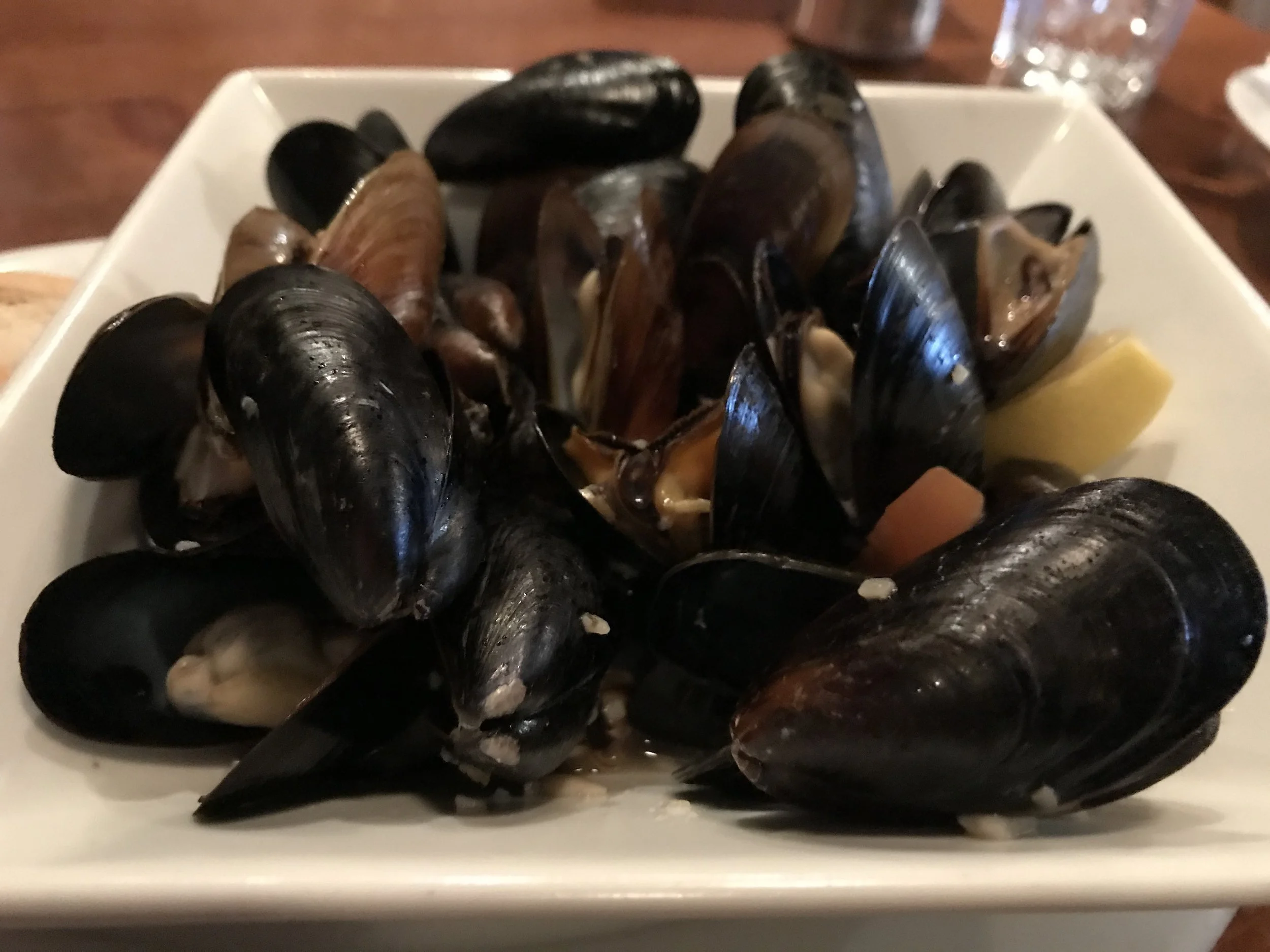 A white square plate filled with cooked mussels garnished with lemon wedges.