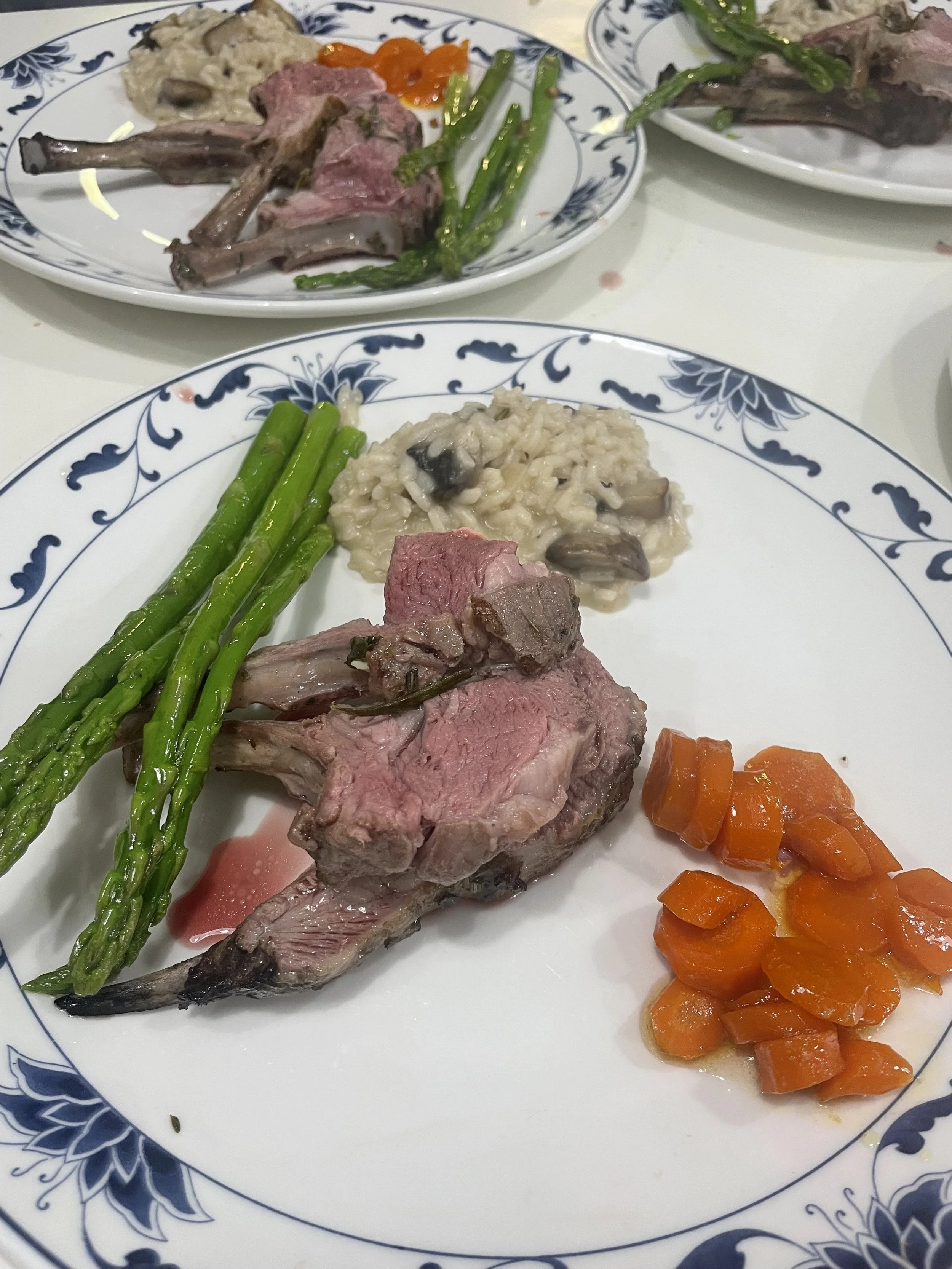 A plate with cooked asparagus, a cut of pink and gray meat, a serving of creamy mushroom risotto, and chopped carrots. In the background, another similar plate with meat, asparagus, and a different side dish.