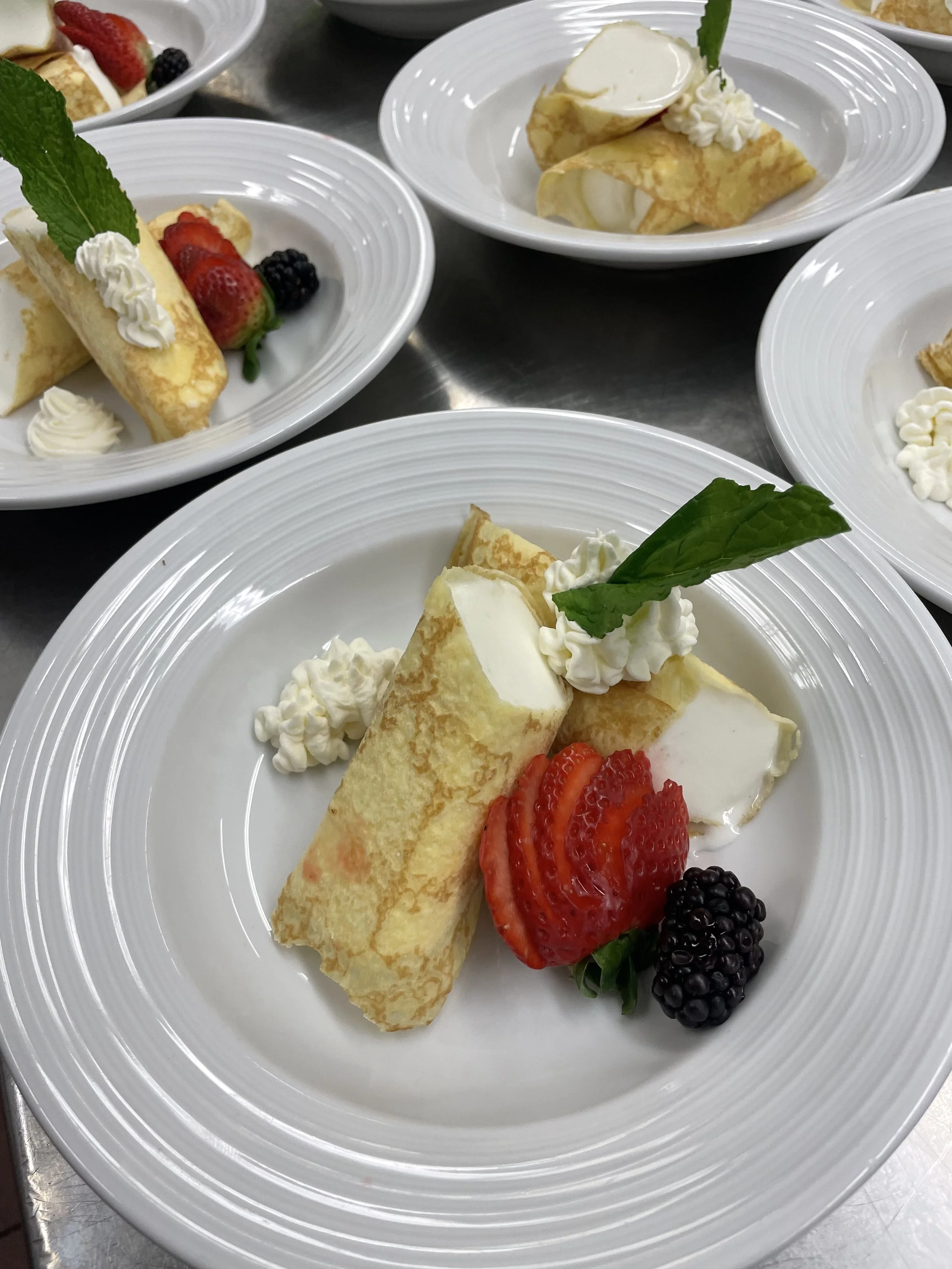 Crepe topped with whipped cream, a strawberry, a blackberry, and a mint leaf, served with additional whipped cream and fruit on a white plate.