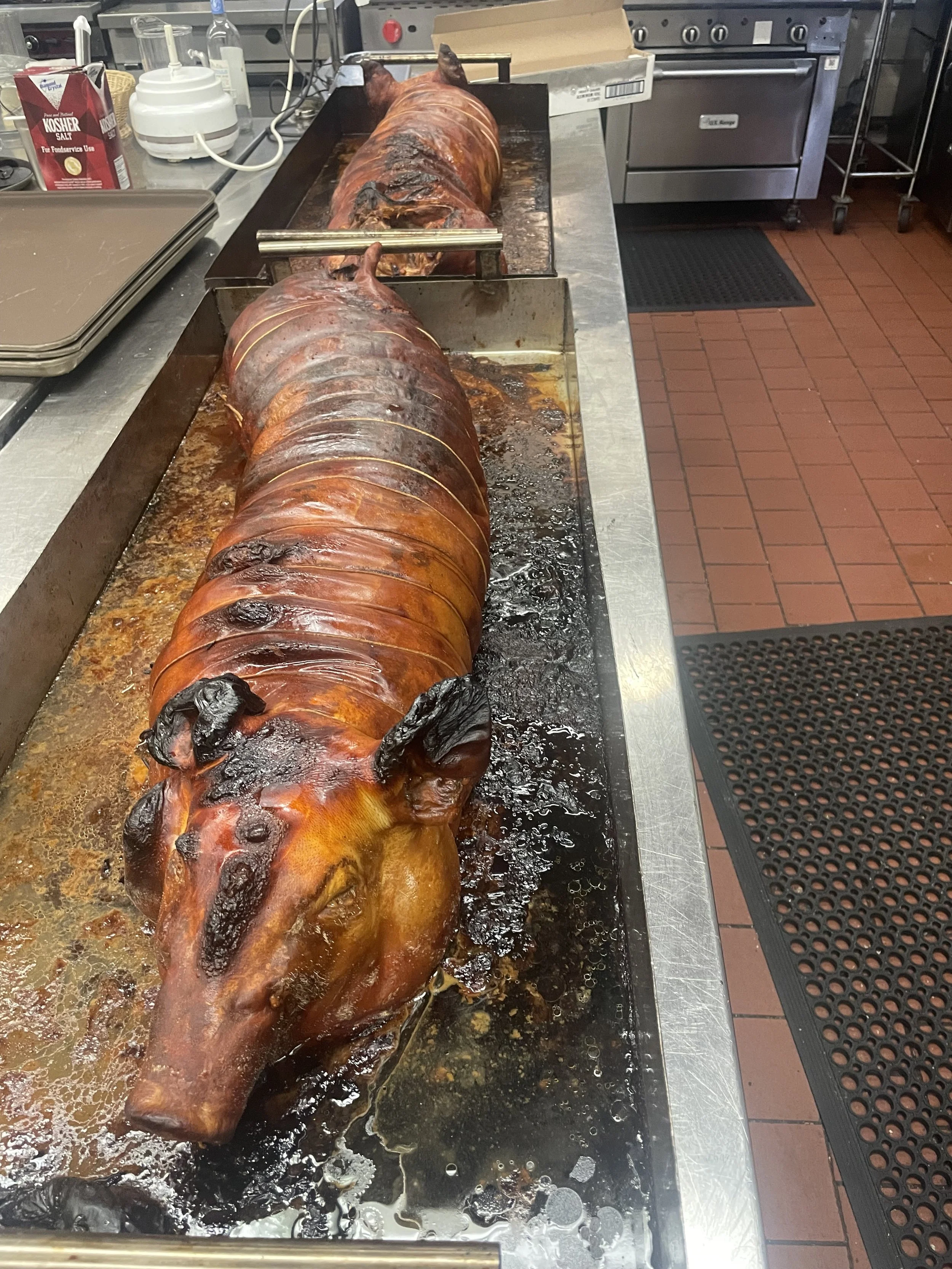 A whole roasted pig on a metal roasting tray in a commercial kitchen.