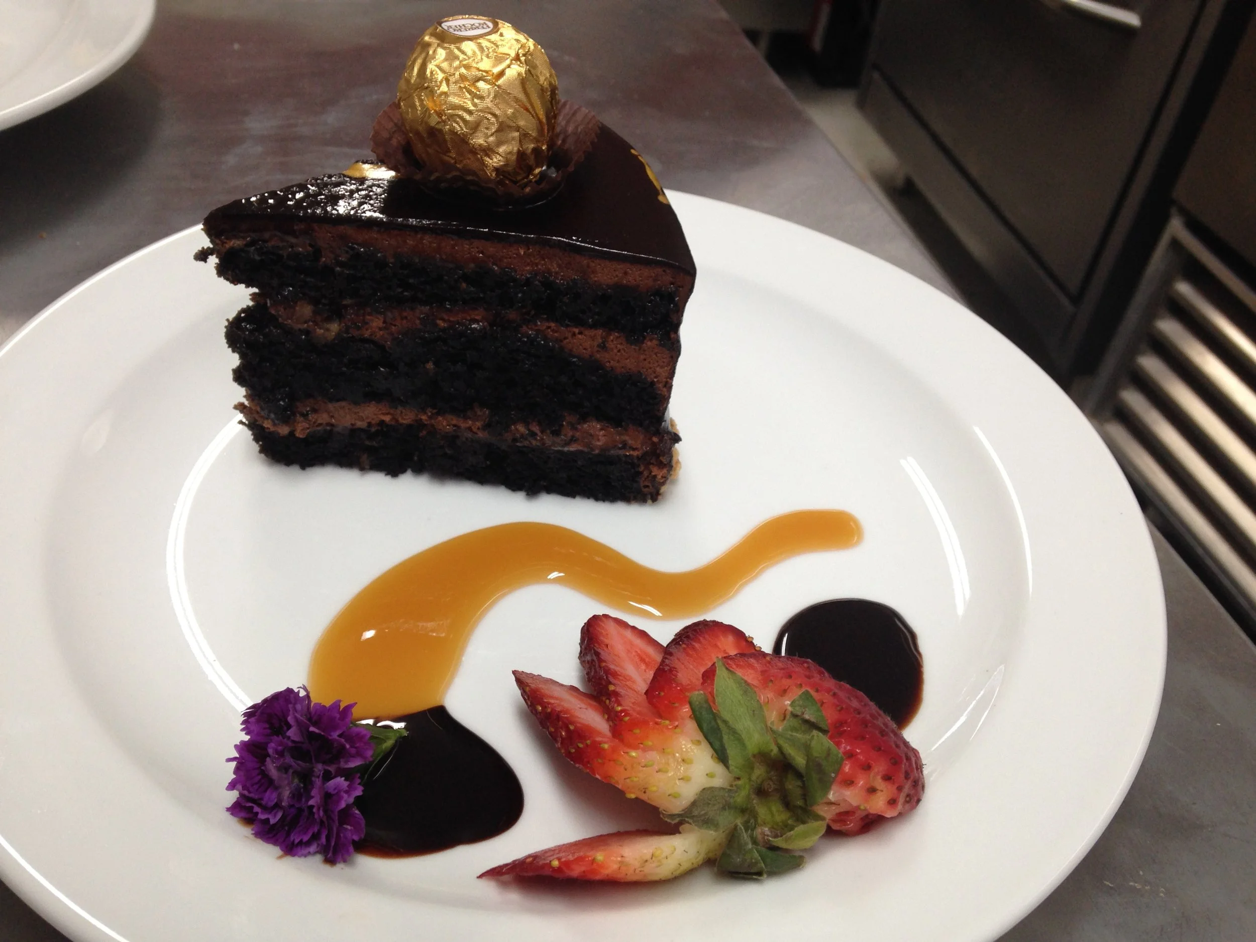 Slice of layered chocolate cake with a chocolate glaze, topped with a gold-wrapped chocolate truffle. Served with sliced strawberries, purple edible flower, and chocolate and caramel sauces on a white plate.
