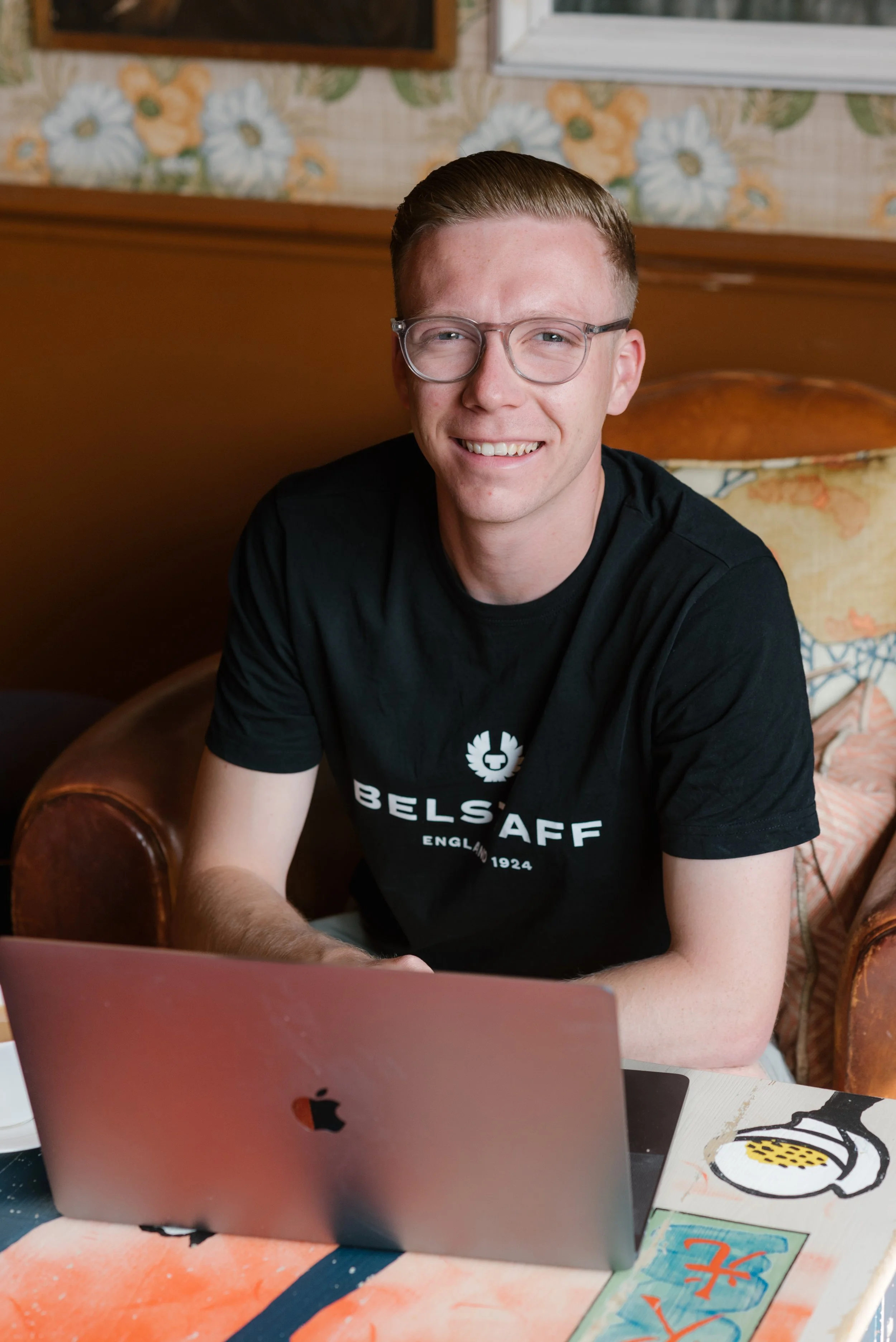 A young man with glasses smiling at the camera, sitting at a table with a laptop, a colorful tablecloth, and artwork, including a plate of fried eggs, in a room with floral wallpaper and a window.