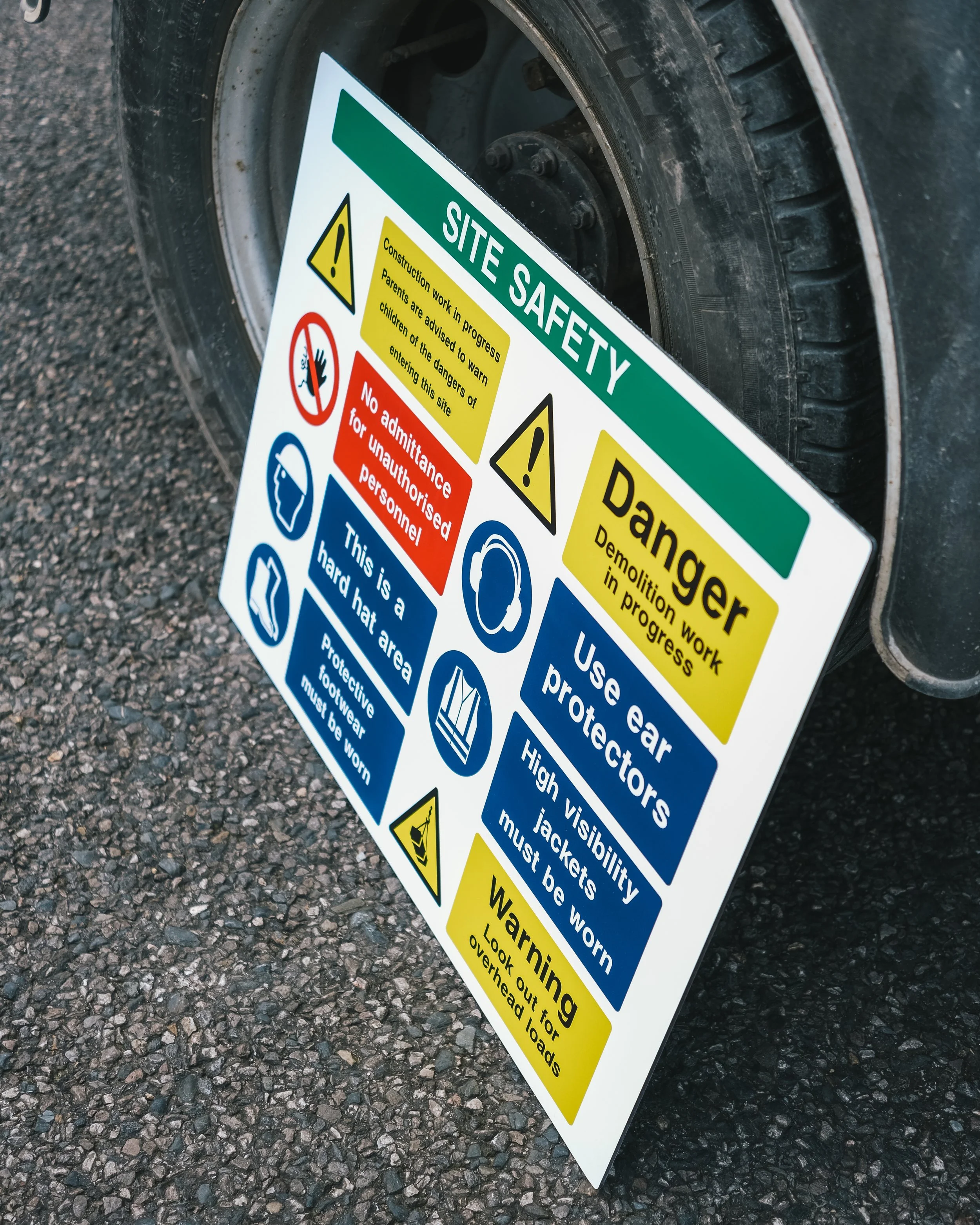 A safety warning sign on a construction site with various warnings about PPE and hazards, leaning against a vehicle tire on a gravel surface.
