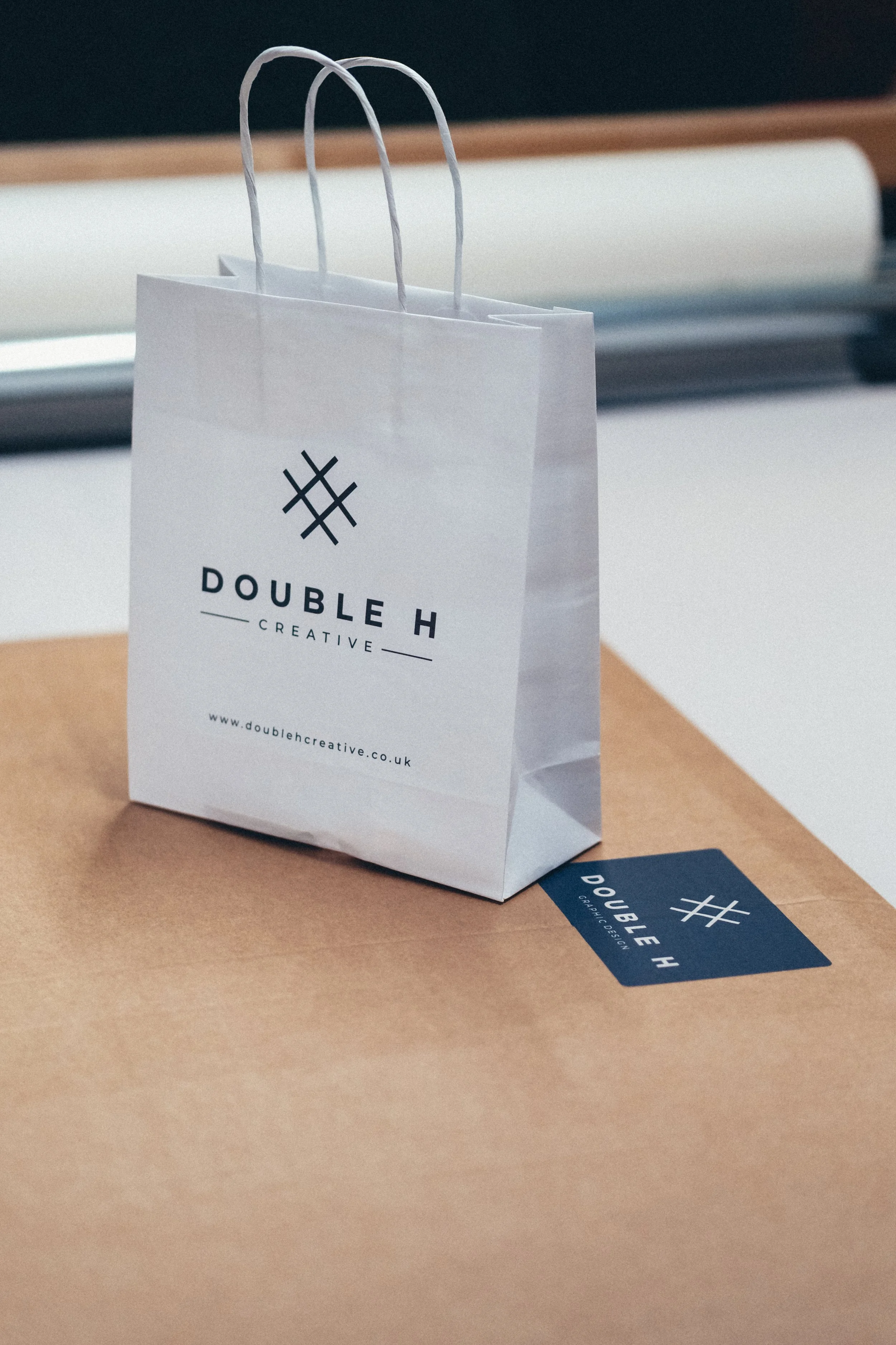 A white paper shopping bag with black logo and text sitting on a light brown surface, with a business card and a black pen nearby. The logo reads 'Double H Creative' and the website is doublehcreative.co.uk.