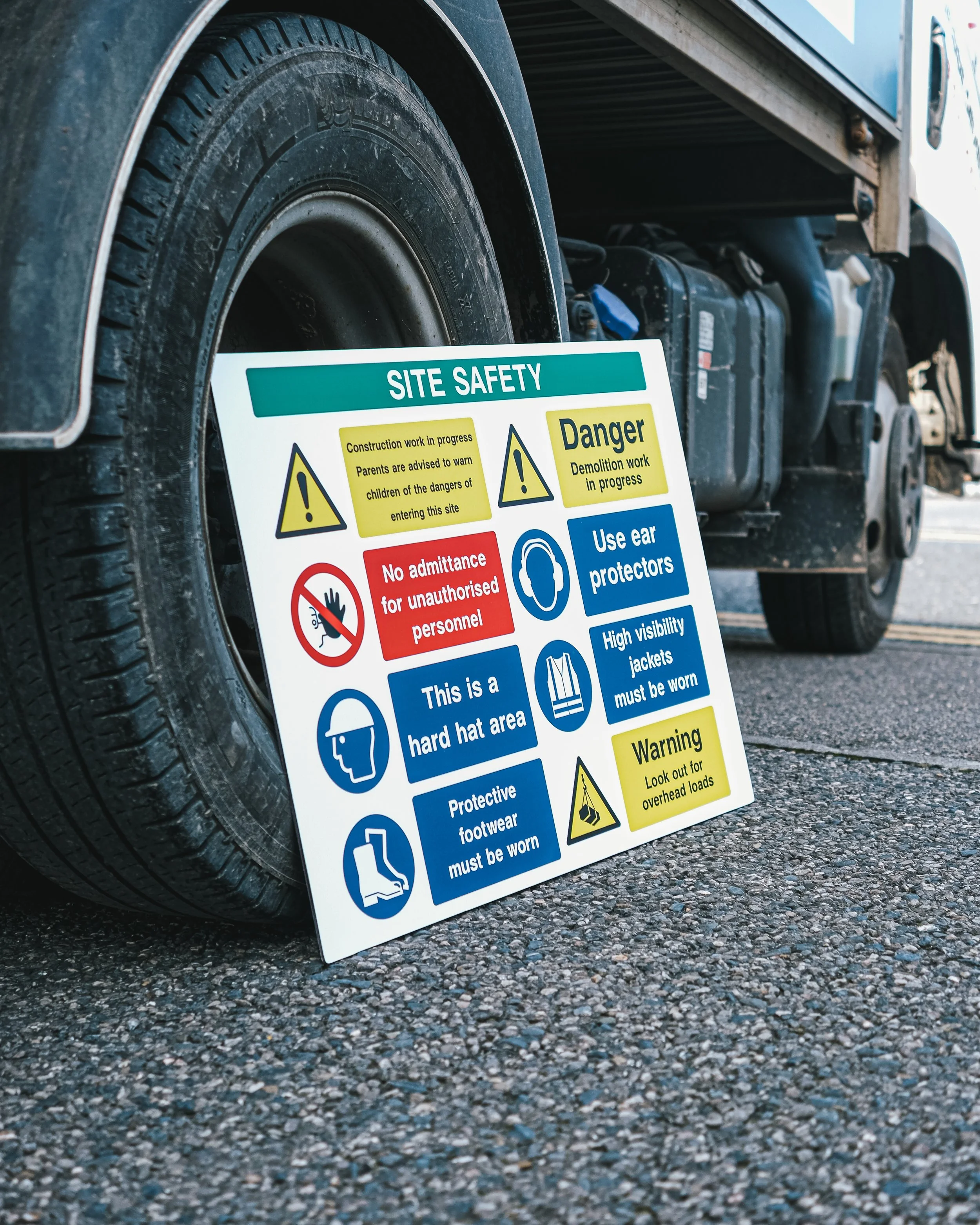 Close-up of a truck tire with a site safety informational sign leaning against it, highlighting safety warnings and instructions for construction and demolition work.
